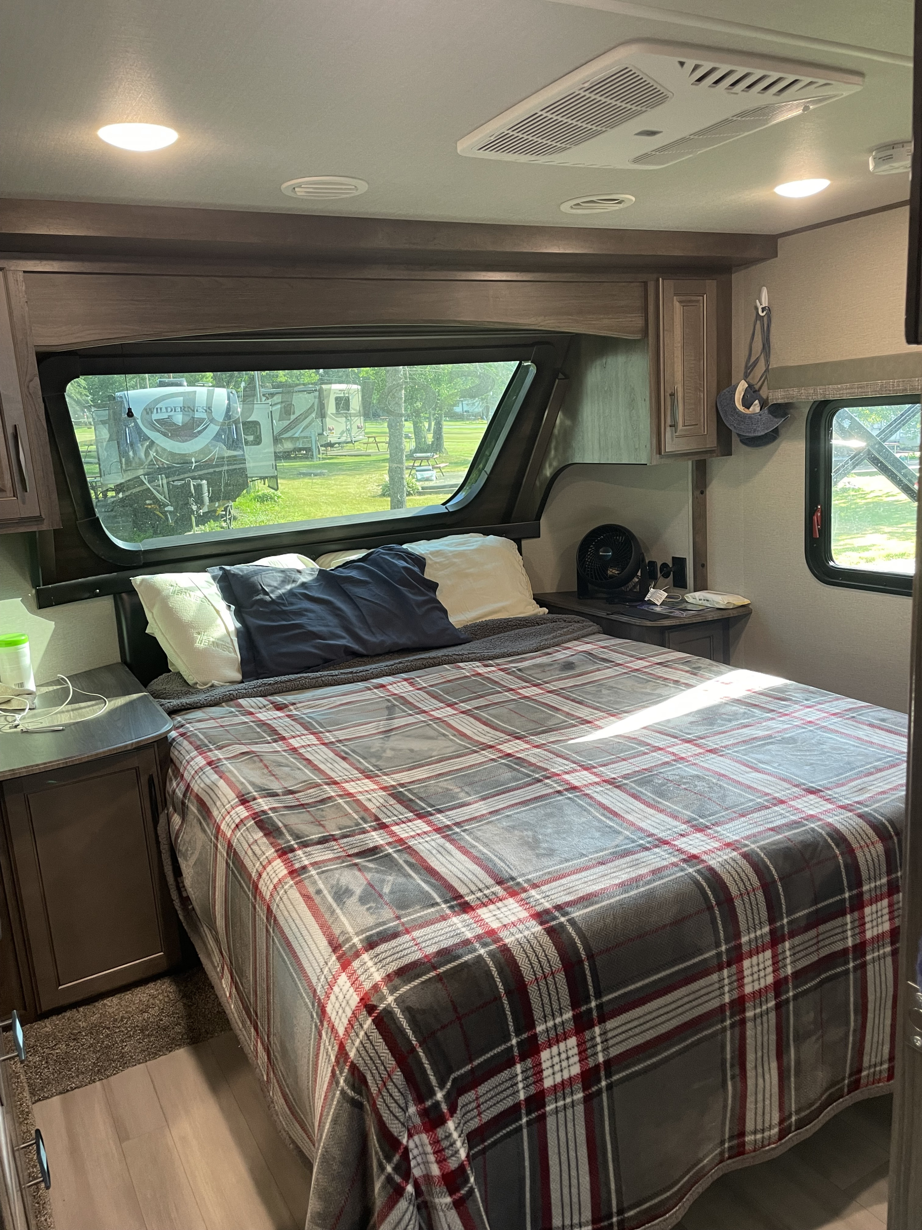 RV rental interior: A cozy bedroom with a plaid comforter, windows revealing other RVs outside, and overhead AC.. Grand Design Solitude S-Class 2021