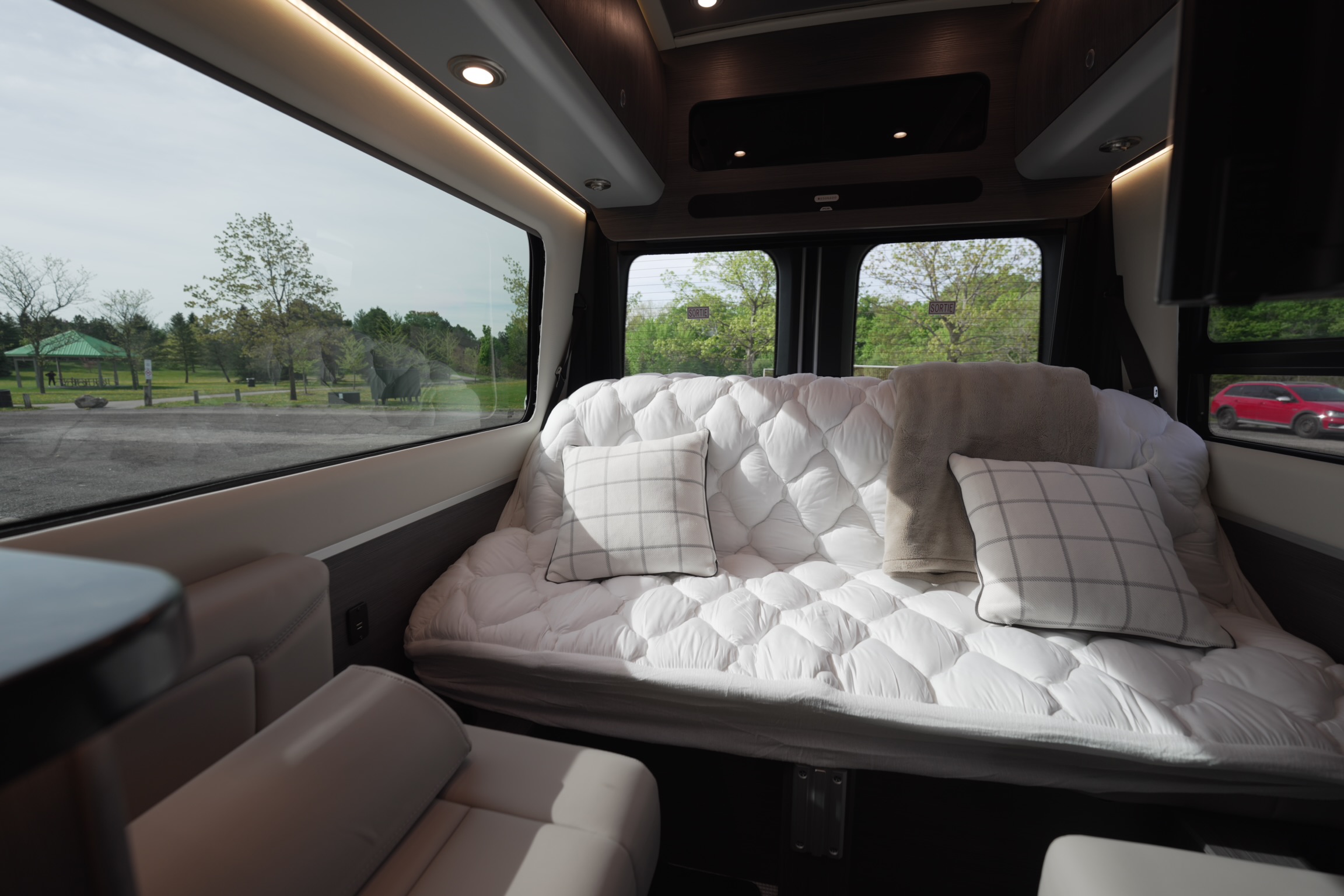 Comfortable RV rental interior featuring a plush white bed, checkered pillows, and a scenic park view through large windows.. Airstream Interstate 2022