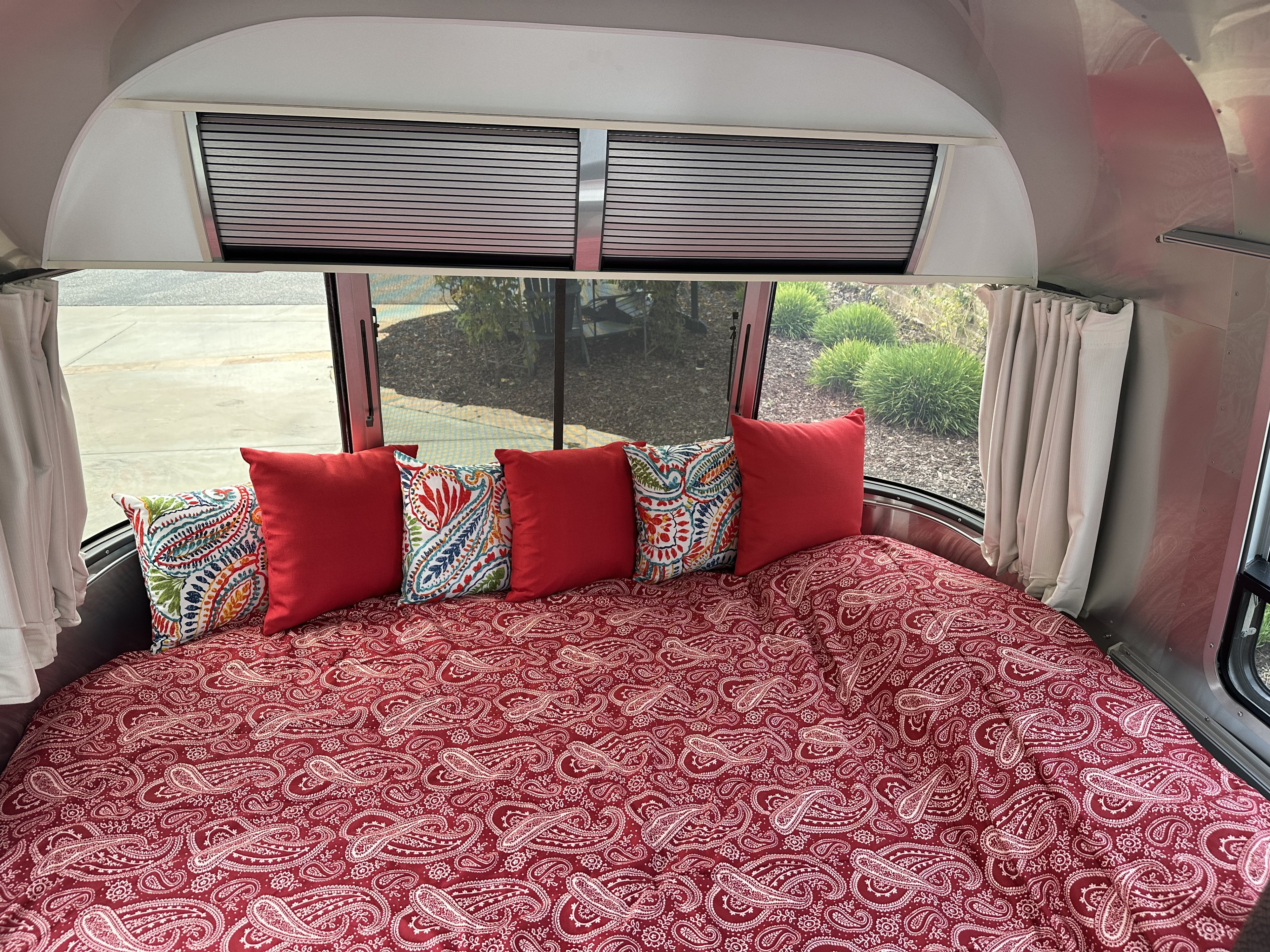 Cozy rv rental bedroom: A comfortable bed with red paisley bedding & colorful pillows in a silver Airstream, with window blinds.. Airstream Sport 2018
