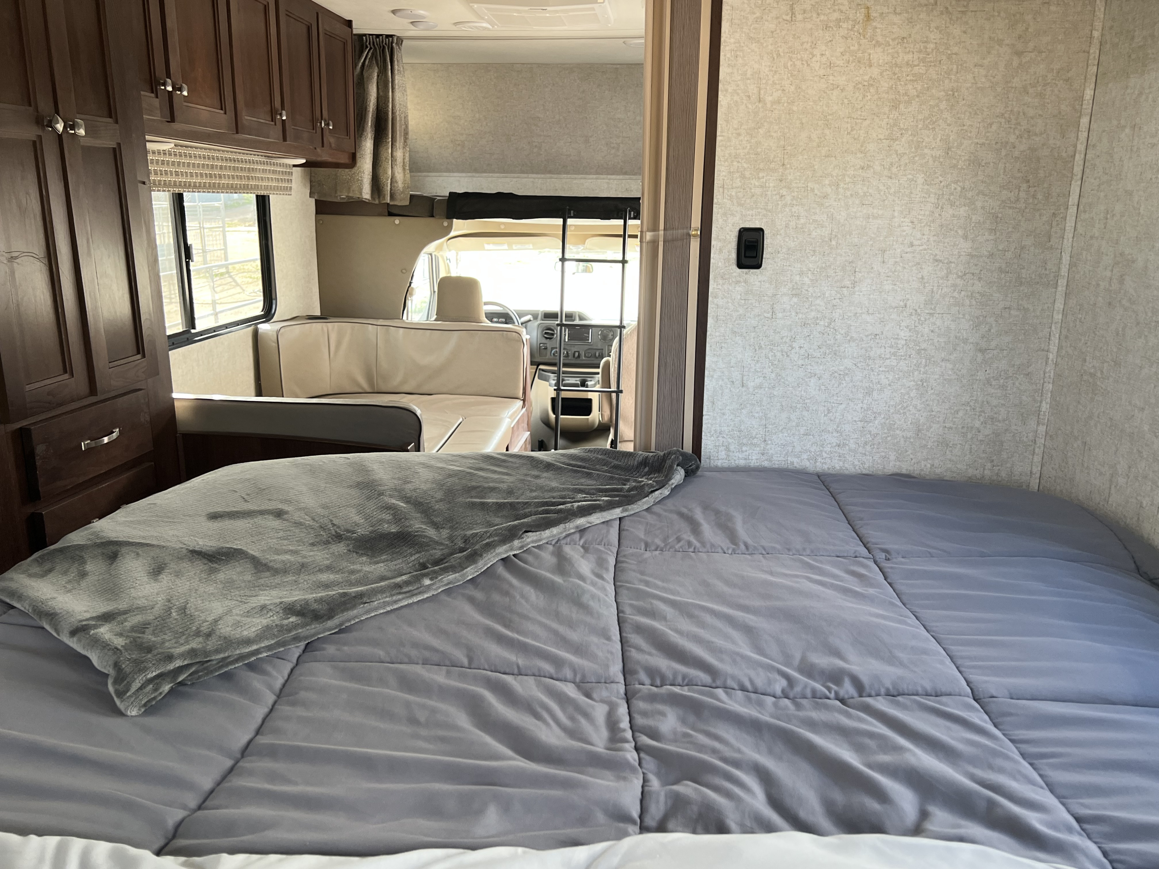 Inside an RV rental, a cozy bed with gray bedding leads to a dining area and the driver's cabin.. Forest River Sunseeker 2019