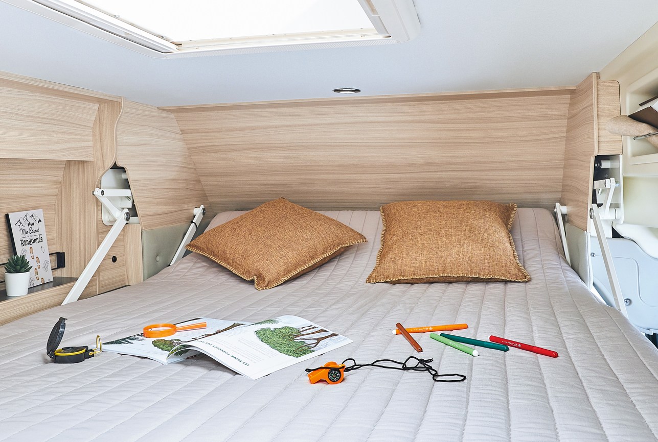 Cozy overhead bed in an RV rental, with light wood interior, skylight, pillows, a book, and compass.. Dreamer Camper Five 2021