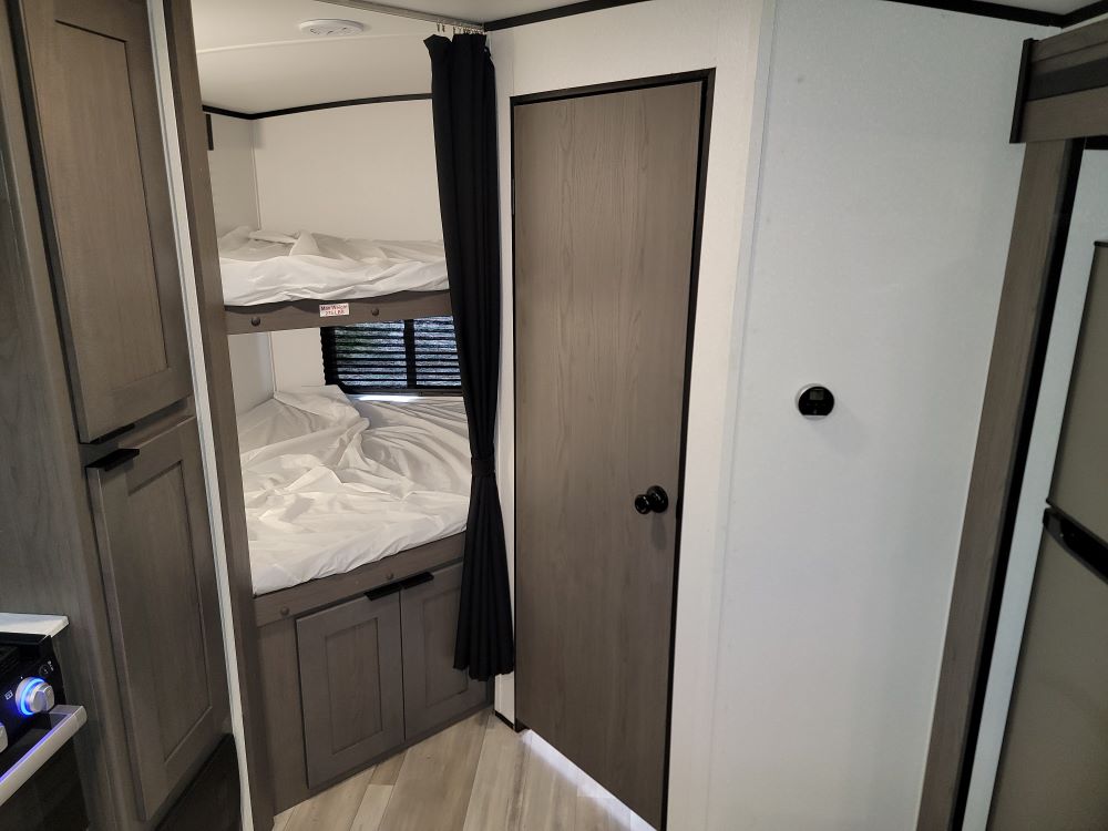 Modern RV rental bunk beds with white linens, black privacy curtain, and wood-toned storage.. Cruiser RV MPG Ultra Lite 2021