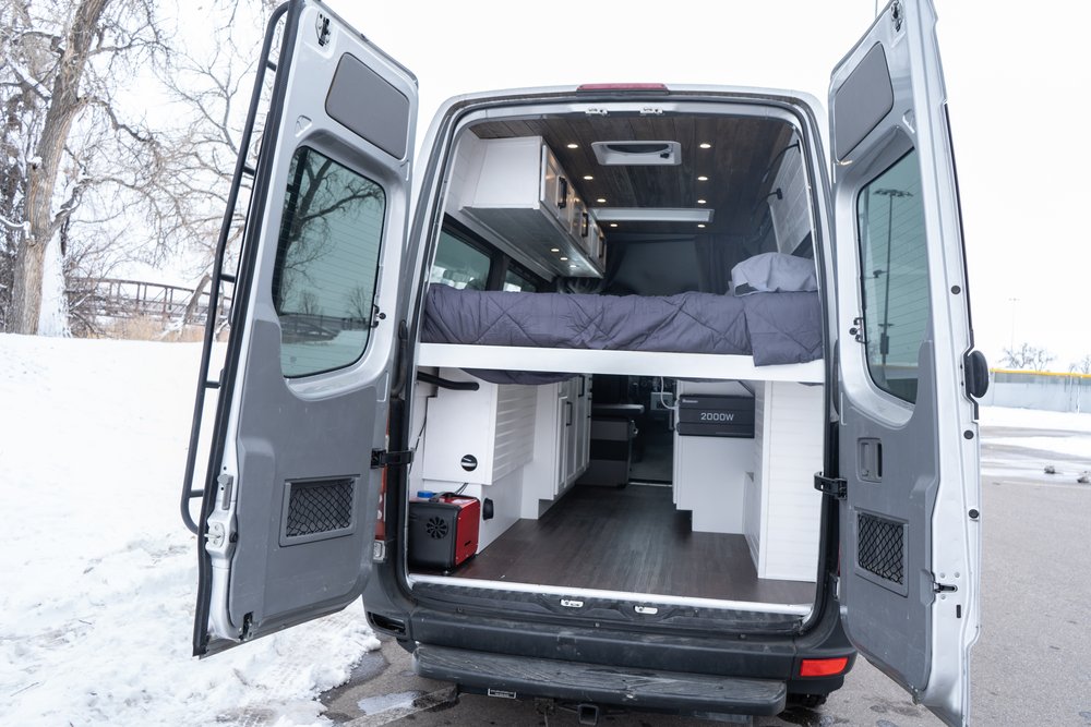 RV rental: Inside a custom camper van with open rear doors, showcasing a raised bed, modern storage, & winter scene.. Mercedes-Benz Sprinter 2011