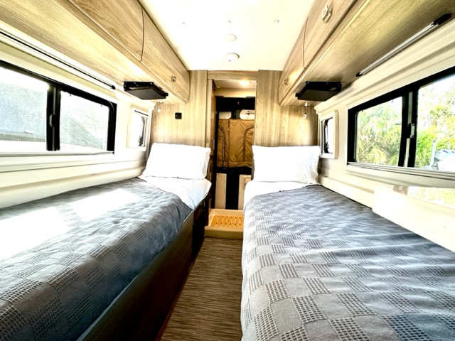 Comfortable twin beds inside an RV rental, complete with overhead cabinets and large windows. Ideal for travel accommodation.. Winnebago Travato 2022