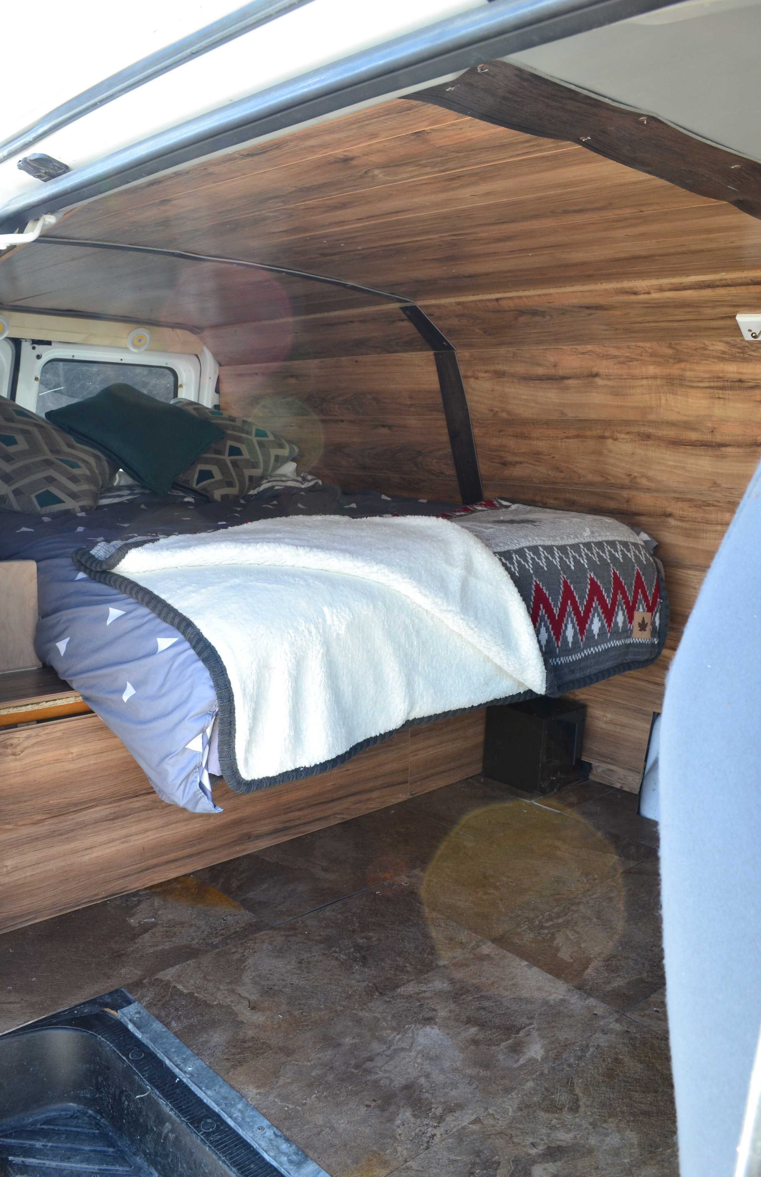 An rv rental's wooden interior, featuring a cozy bed with blankets and pillows, ideal for comfortable camper van trips.. Glacier Campers #Vanlife Camper 2014