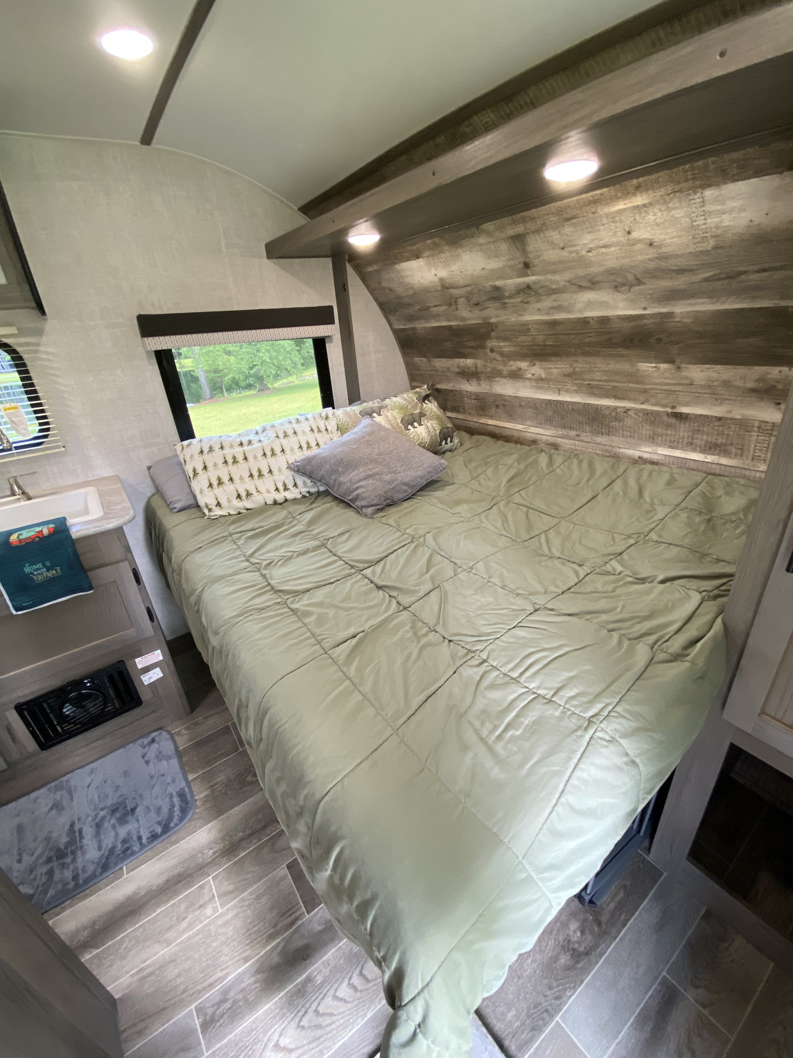 RV rental interior: Cozy bedroom featuring a green bed, rustic wood-look wall, window view, and a compact sink area.. Gulf Stream Ameri-Lite Super Lite 2021