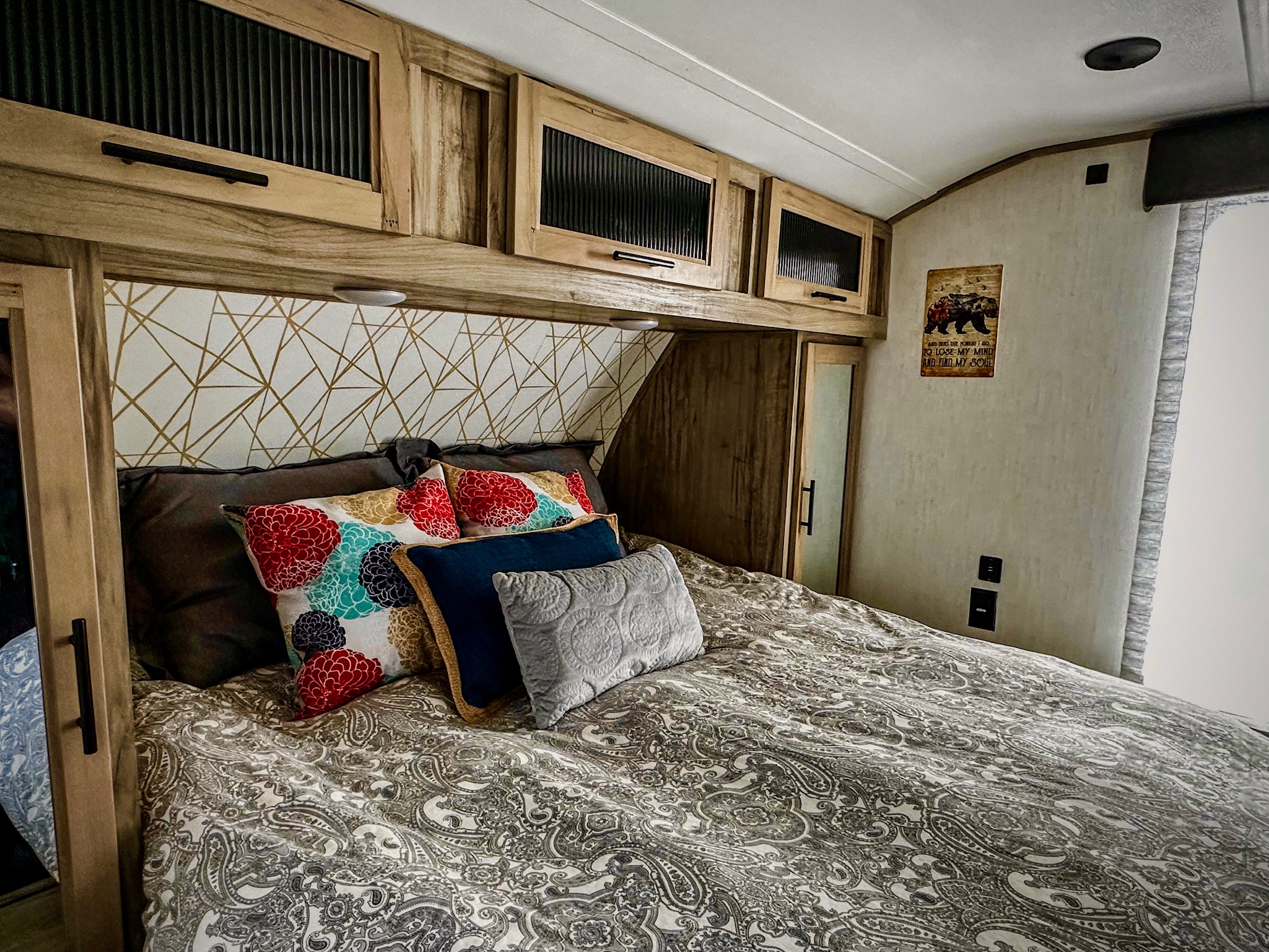 Inviting RV rental bedroom: bed with paisley comforter, colorful pillows, geometric accent wall, and overhead storage. Bear sign on wall.. Forest River Cherokee Alpha Wolf 2021