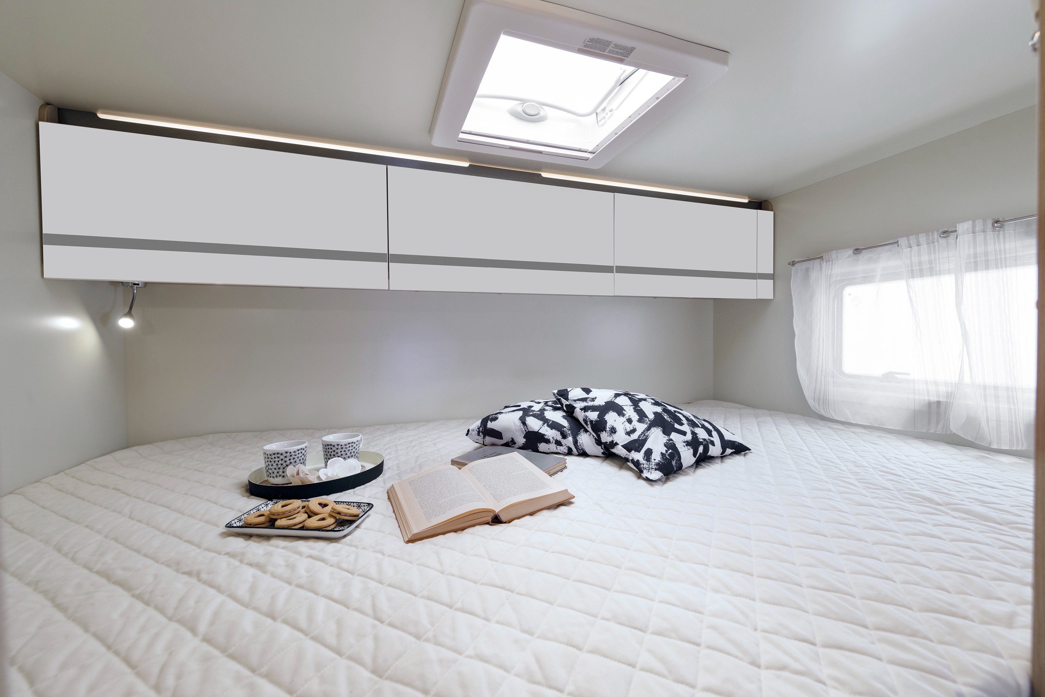 Cozy RV rental bedroom with bright skylight, white bed, overhead cabinets, and a tray with snacks and a book.. Giottiline Siena 440 2023