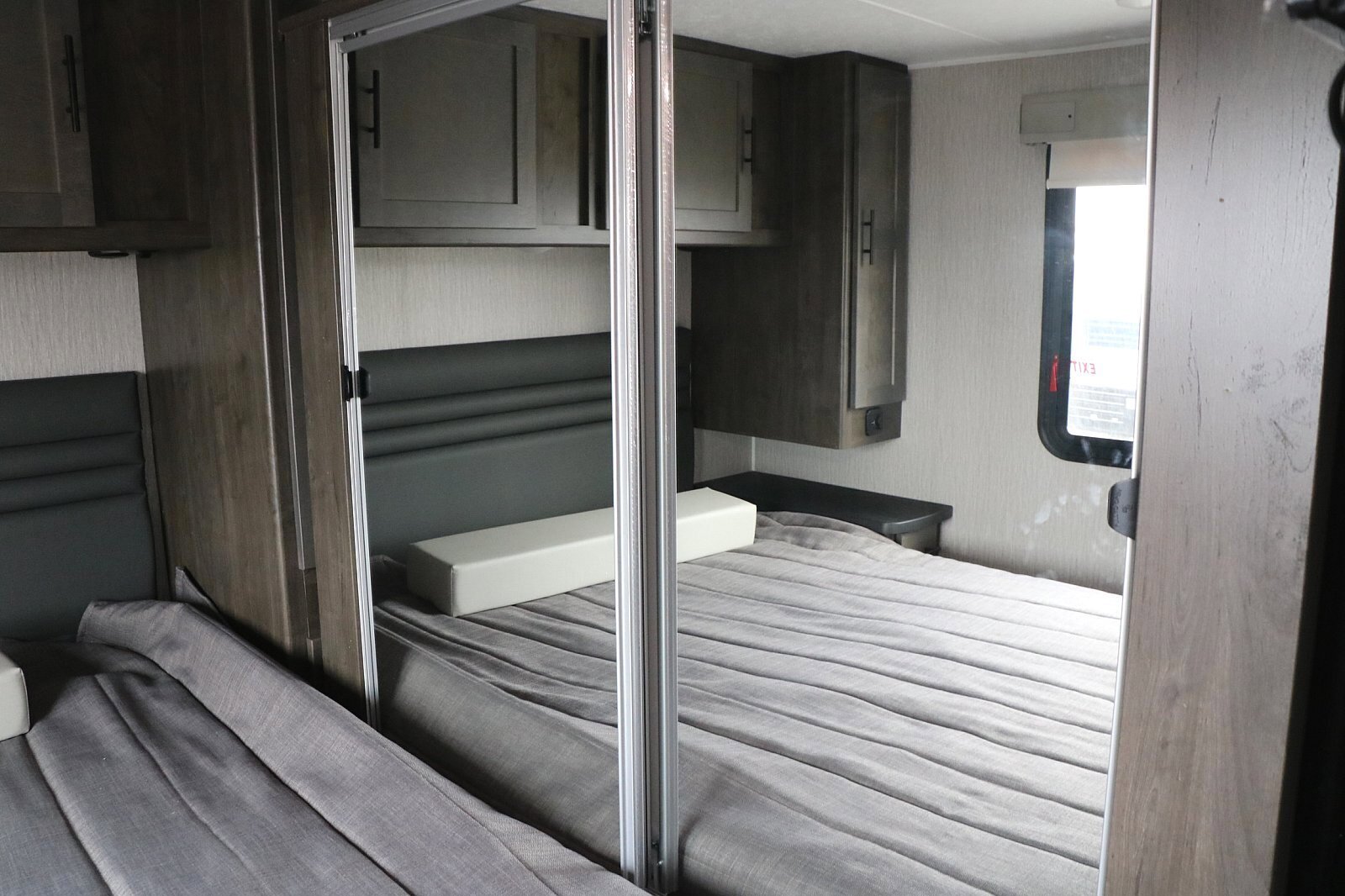 Inside an RV rental bedroom: two beds with grey linens, overhead storage, and a central mirrored partition.. Coachmen Leprechaun 2022