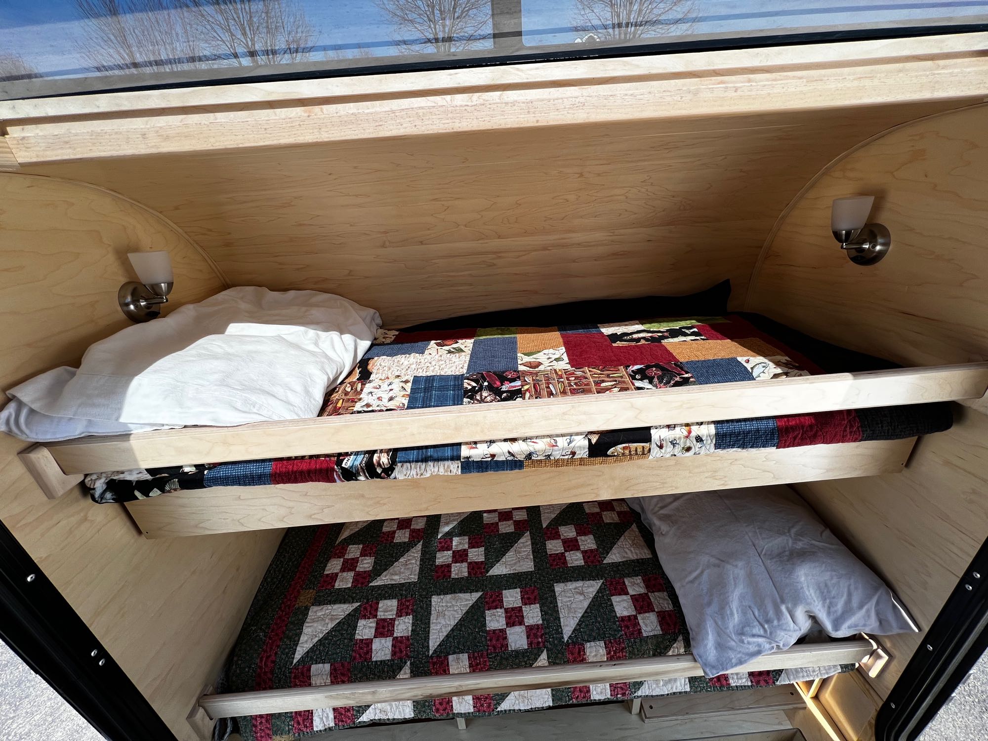 Inside an RV rental: two wooden bunk beds with patterned quilts, pillows, and wall-mounted lights for comfort.. Colorado Teardrop Trailer Summit 2022