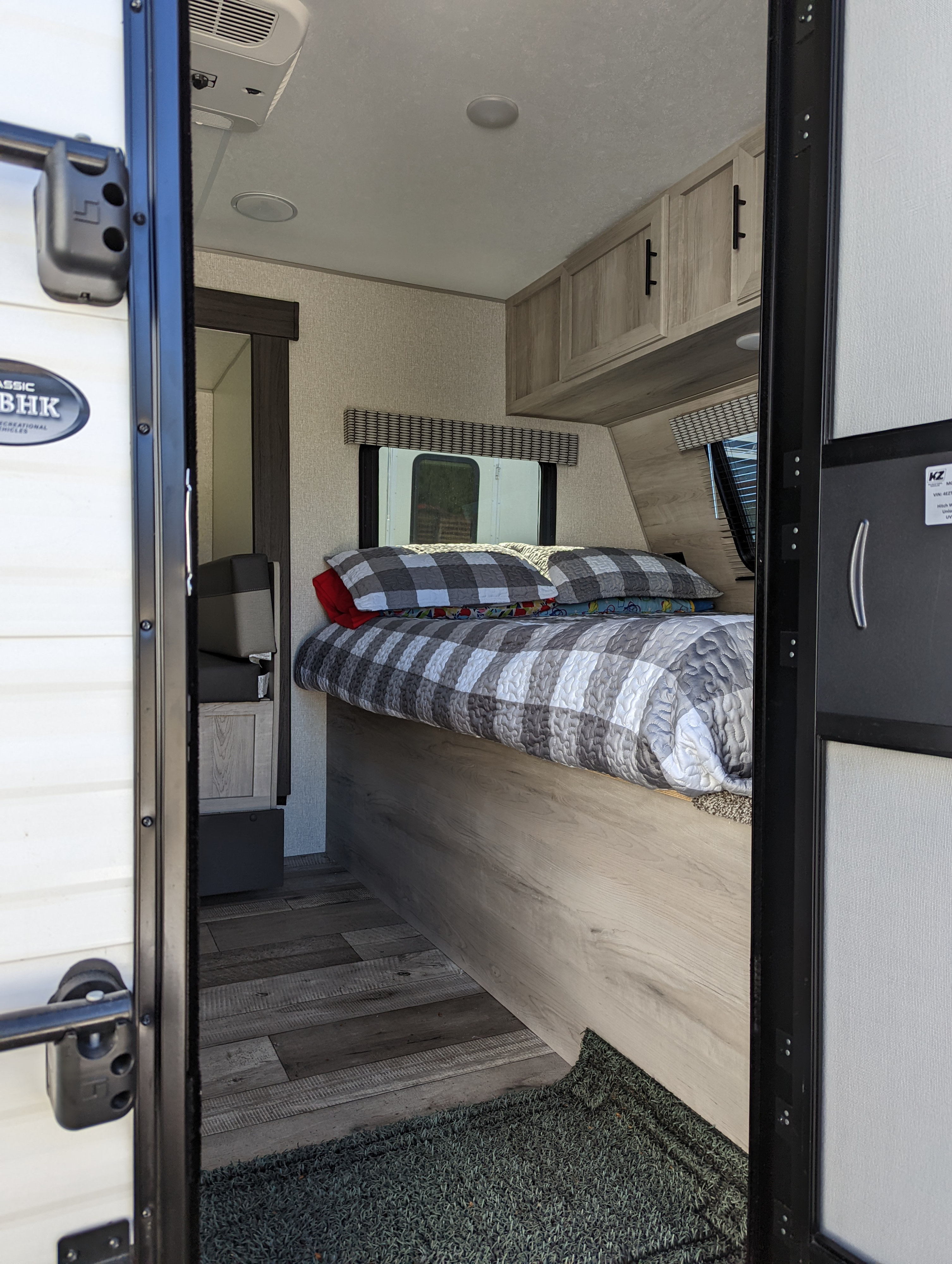 Cozy RV rental bedroom featuring a comfortable bed with checkered bedding, overhead cabinets, and modern wood-look flooring.. KZ 191BHK 2022