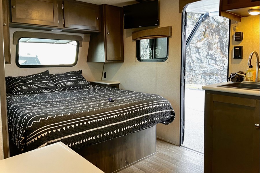 RV rental interior: cozy bedroom with patterned bed & overhead cabinets, plus a compact kitchen area & outdoor view.. Forest River Viking 2020