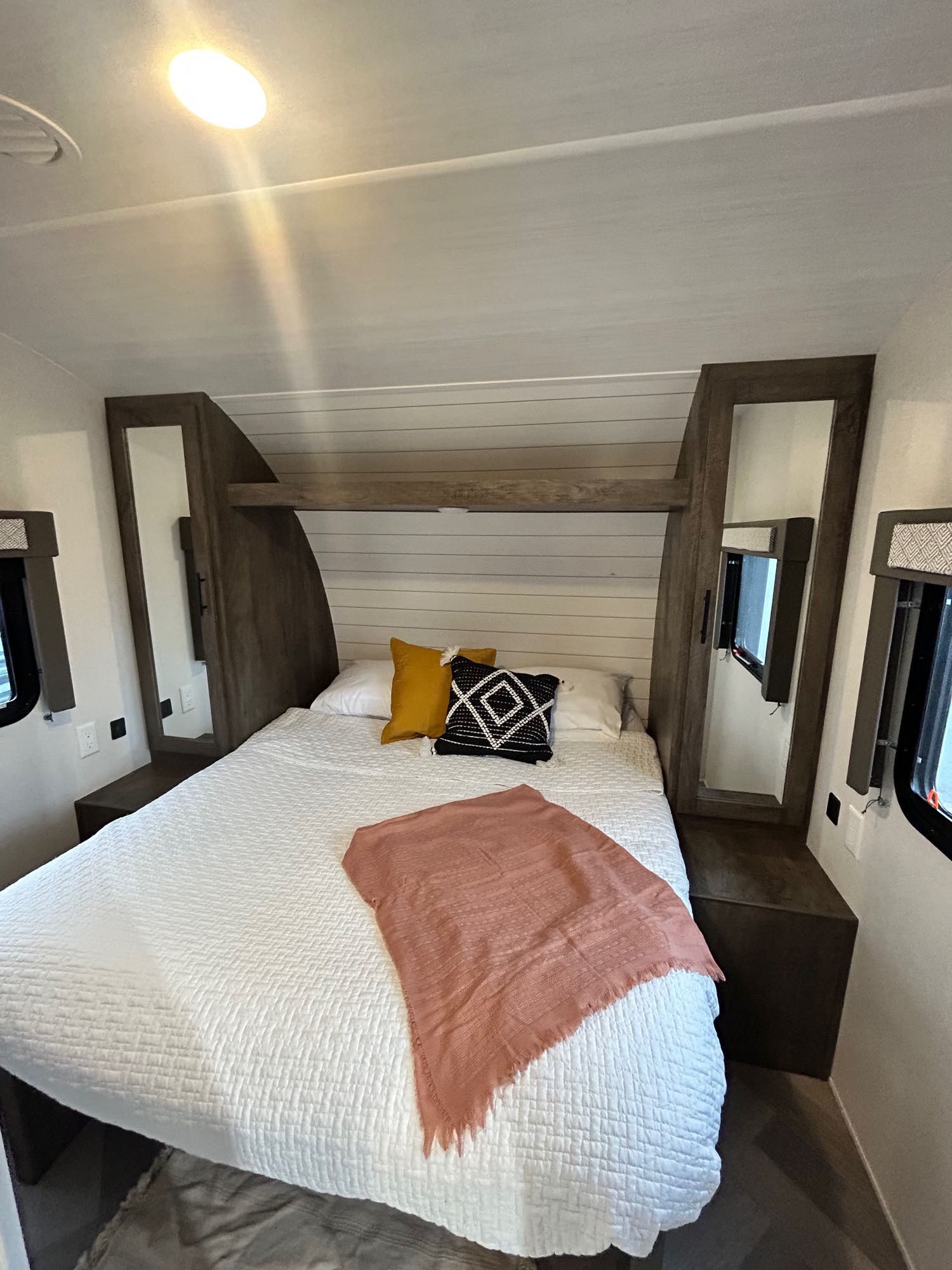 Comfortable RV rental bedroom featuring a white bedspread, pink throw, and dark wood built-ins with mirrors. Ideal for travel.. Forest River Wildwood X-Lite 2021