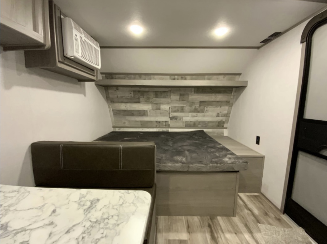 Cozy RV rental interior showing a bed area with a wood-look headboard, an overhead AC unit, and a dinette table/bench.. Coleman Lantern 2023