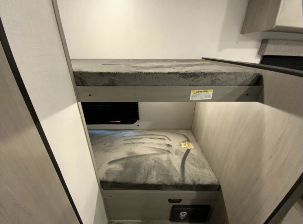 Inside an RV rental: comfortable stacked bunk beds with soft grey bedding, featuring a caution label and a vent under the top bunk.. Coleman Lantern 2023