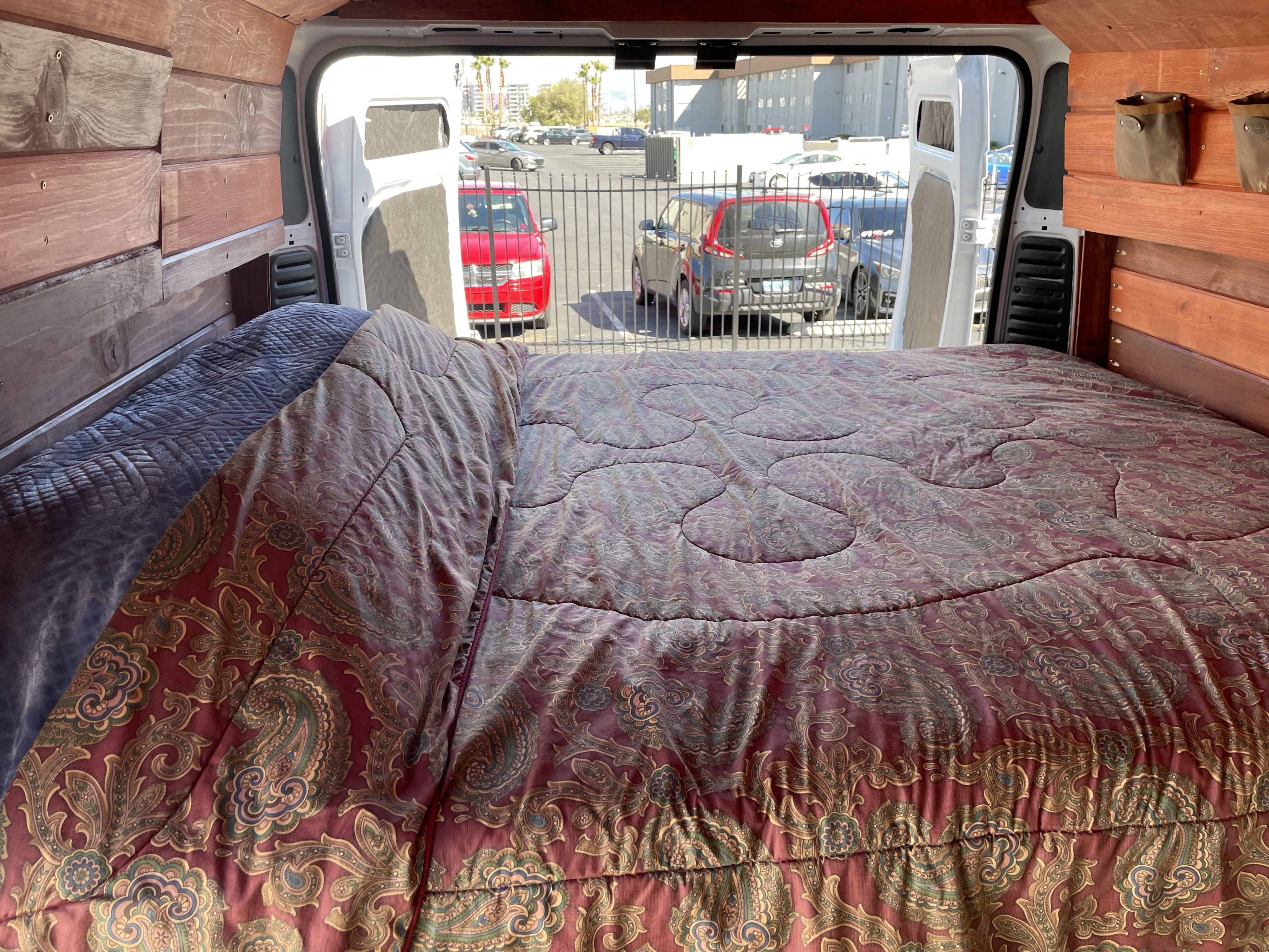 Cozy RV rental camper van interior featuring a large bed, wood-paneled walls, and a view of a sunny parking lot.. Dodge Other 2021