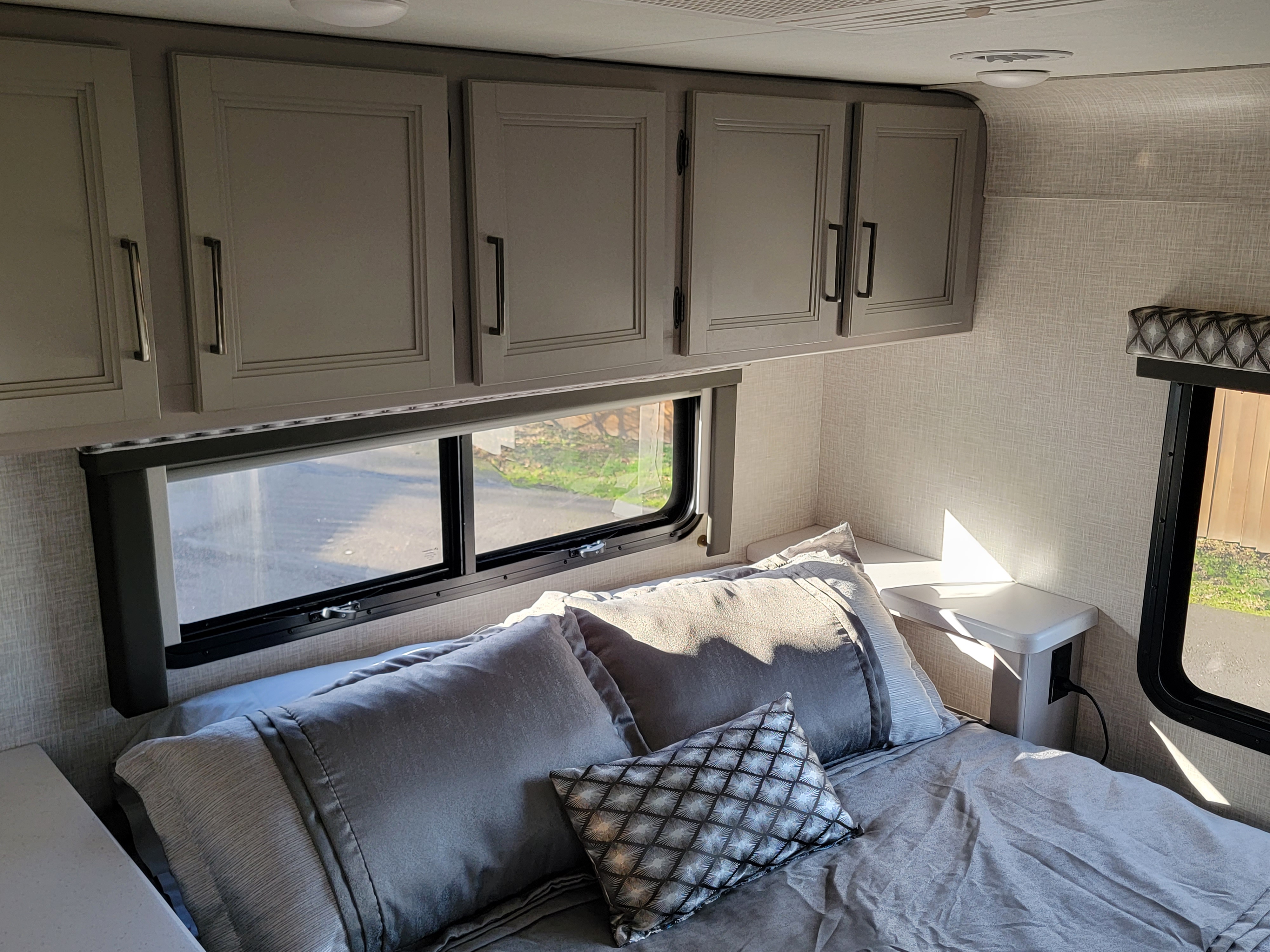 Modern RV rental bedroom featuring ample overhead cabinets, a window, and a comfy bed with side table.. Thor Motor Coach Quantum 2023