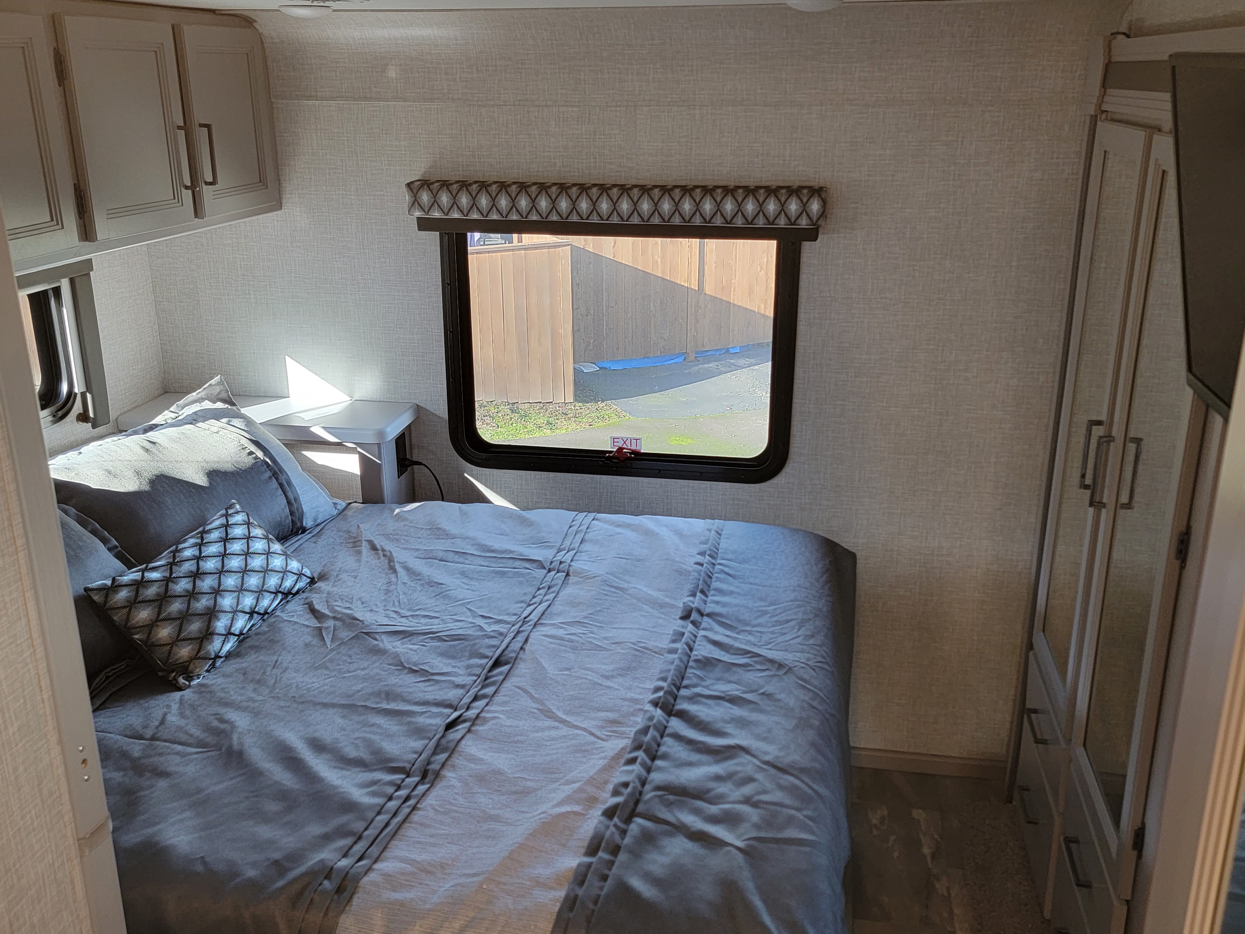 Cozy RV rental bedroom featuring a comfy bed with grey bedding, overhead cabinets, a tall mirrored closet, and a sunlit window.. Thor Motor Coach Quantum 2023