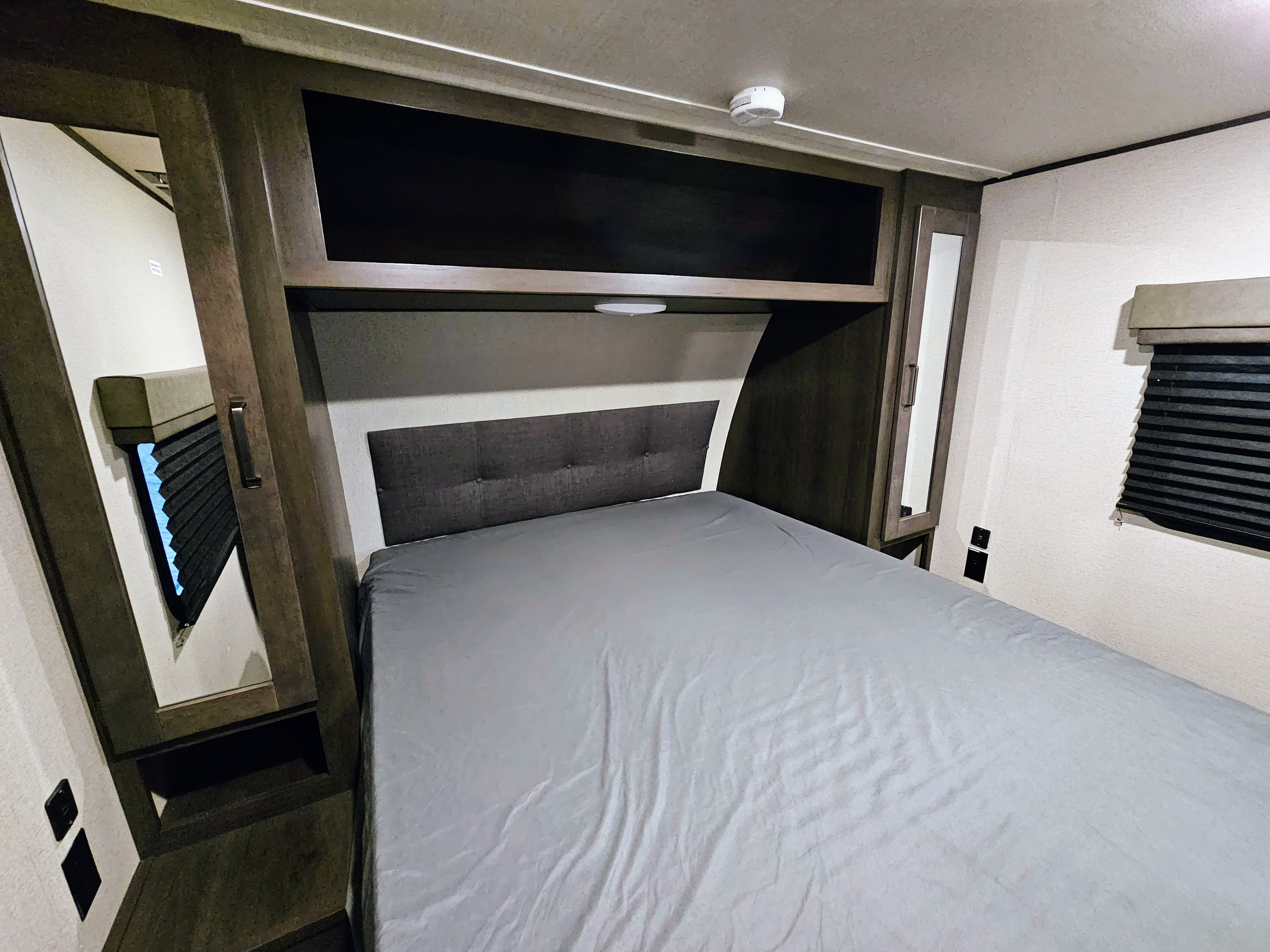 Modern RV rental bedroom with a grey bed, tufted headboard, overhead storage, and mirrored closets. Includes window and outlets.. Grand Design Transcend 2022