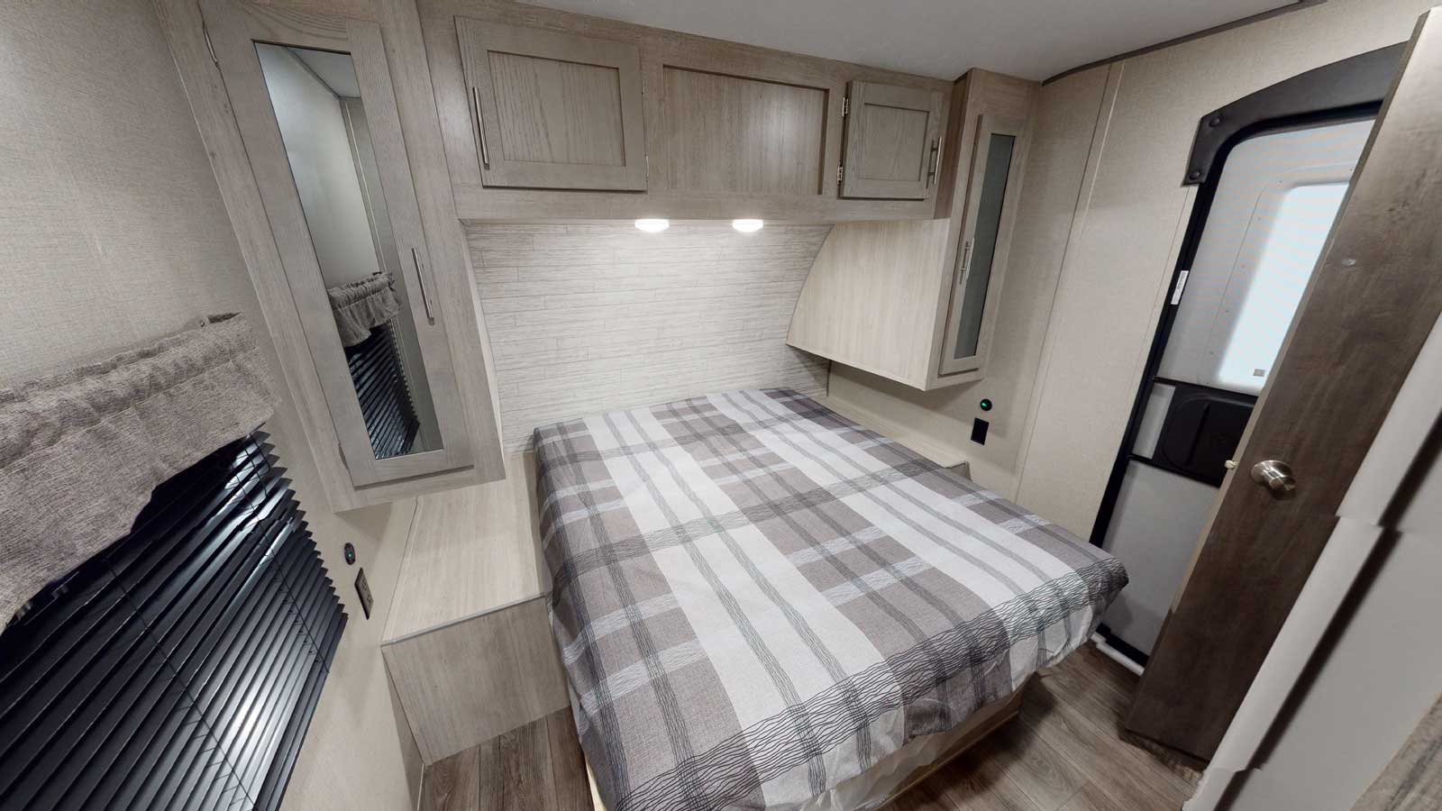 Light-toned RV rental bedroom with plaid bedding, overhead cabinets, and window blinds for a cozy stay.. Forest River Catalina legacy 2023