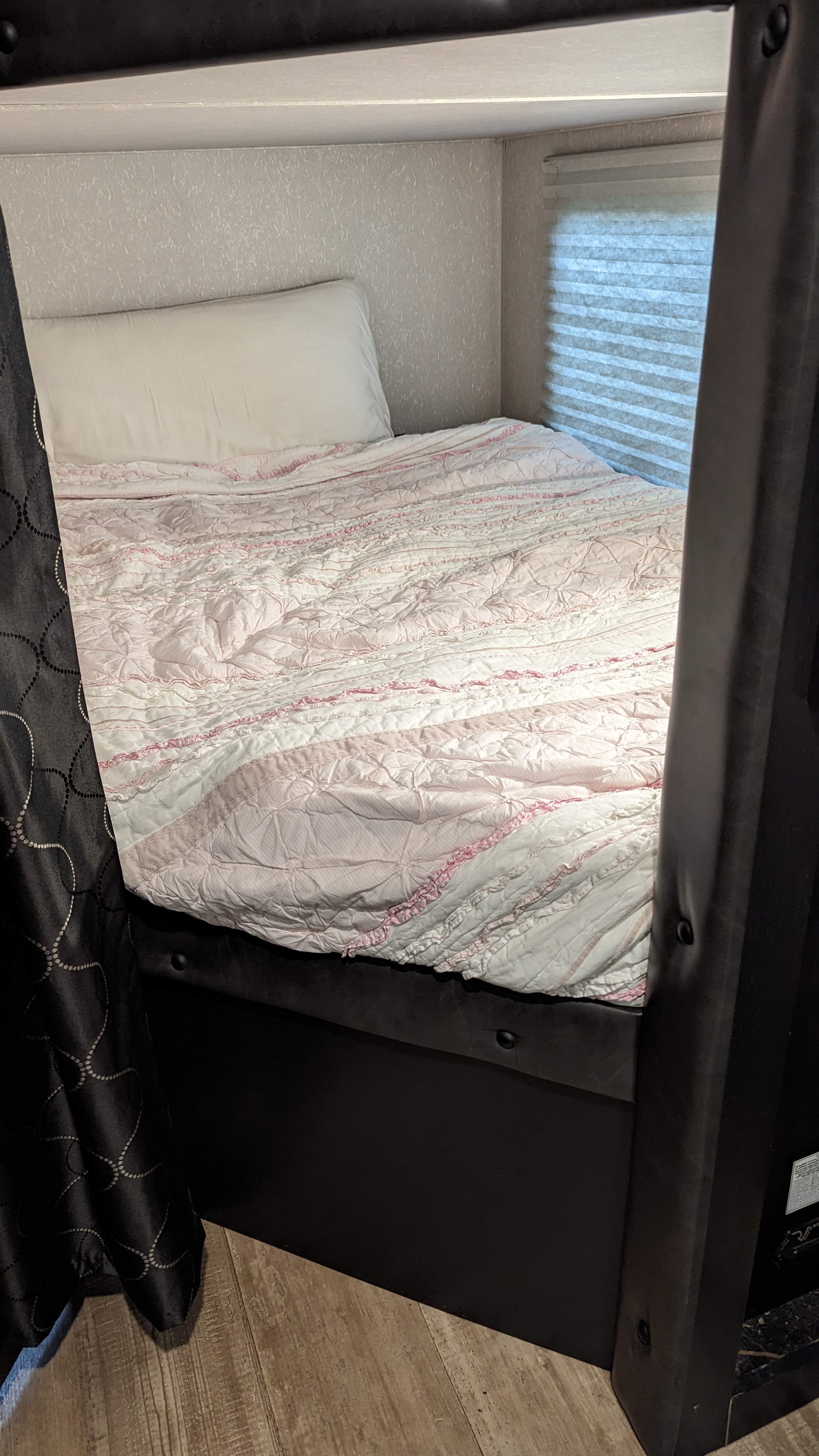 This RV rental features a cozy bunk bed with pink and white ruffled bedding, a white pillow, and window blinds, set in a compact space.. Forest River Cherokee Grey Wolf Black Label 2021