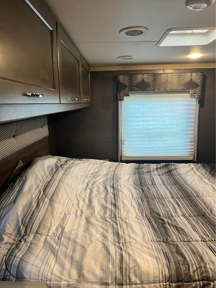 Cozy RV rental bedroom featuring a striped bed, overhead cabinets, and a window with blinds, ready for your next adventure.. Winnebago Minnie Winnie 2017