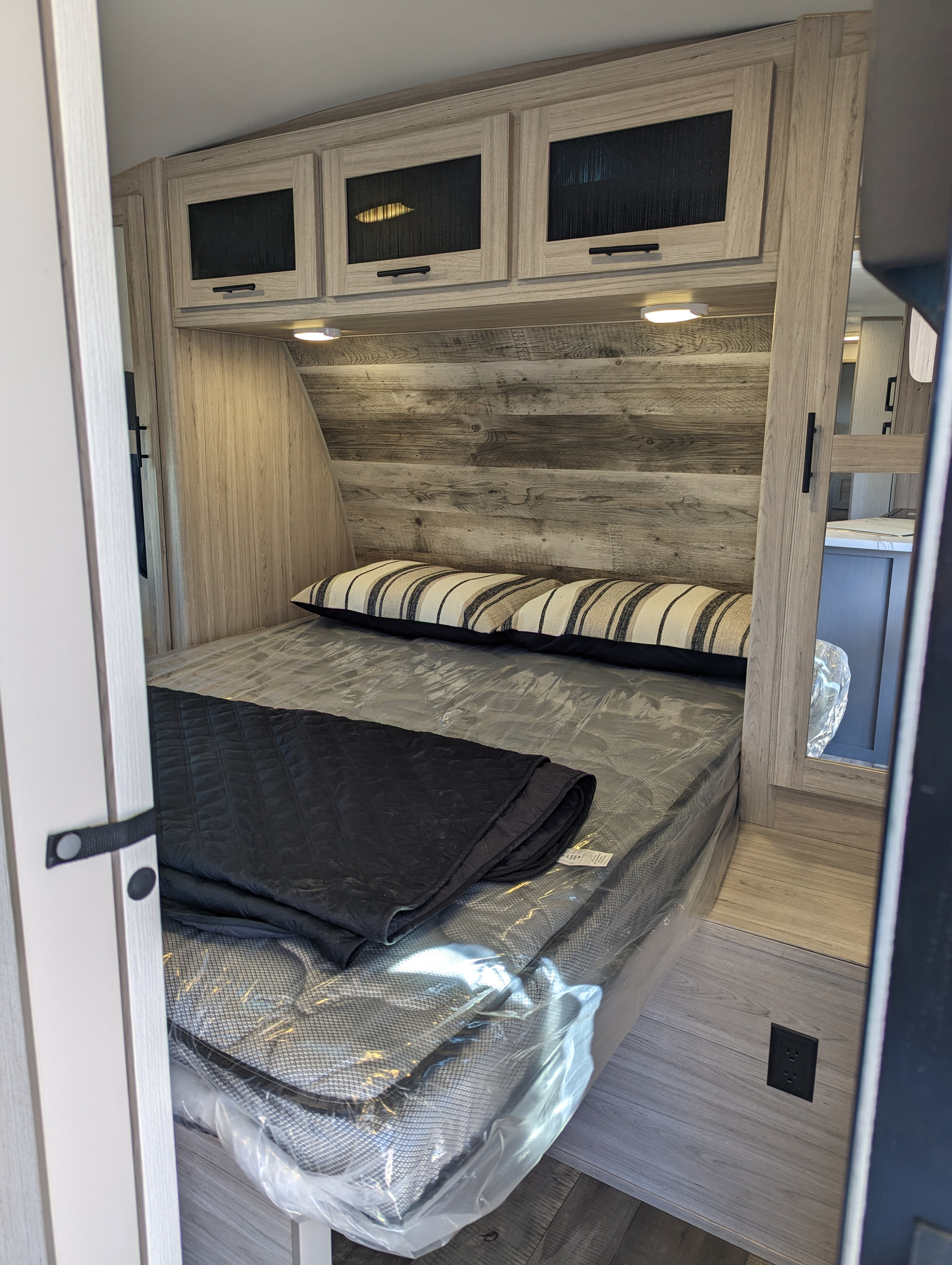 Inside a modern RV bedroom, ready for your rv rental. Features a bed with plastic-wrapped mattress, overhead cabinets, and wood-paneled wall.. Heartland Sundance 2022
