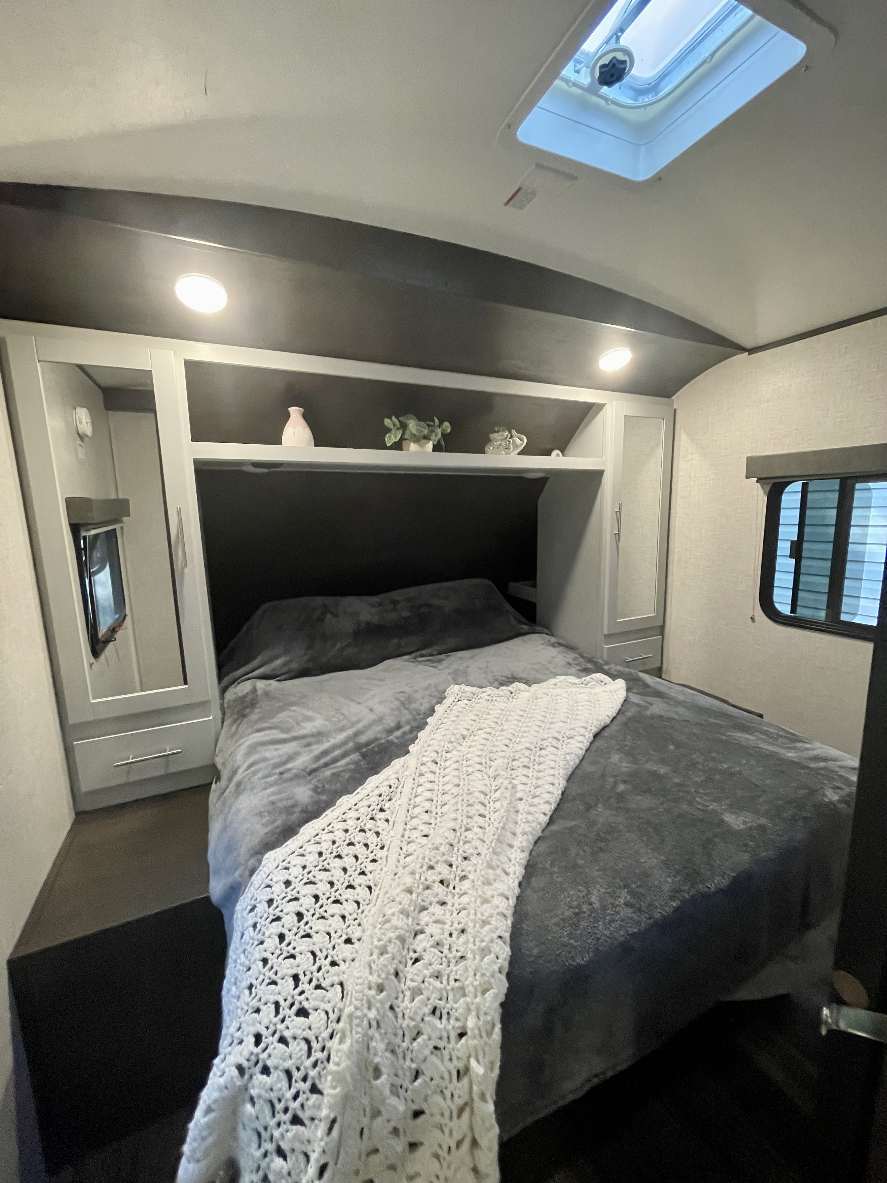 Full view of a modern RV rental bedroom featuring a comfy grey bed, white crochet throw, overhead shelving, and side window.. Keystone RV Bullet Premier Ultra 2020