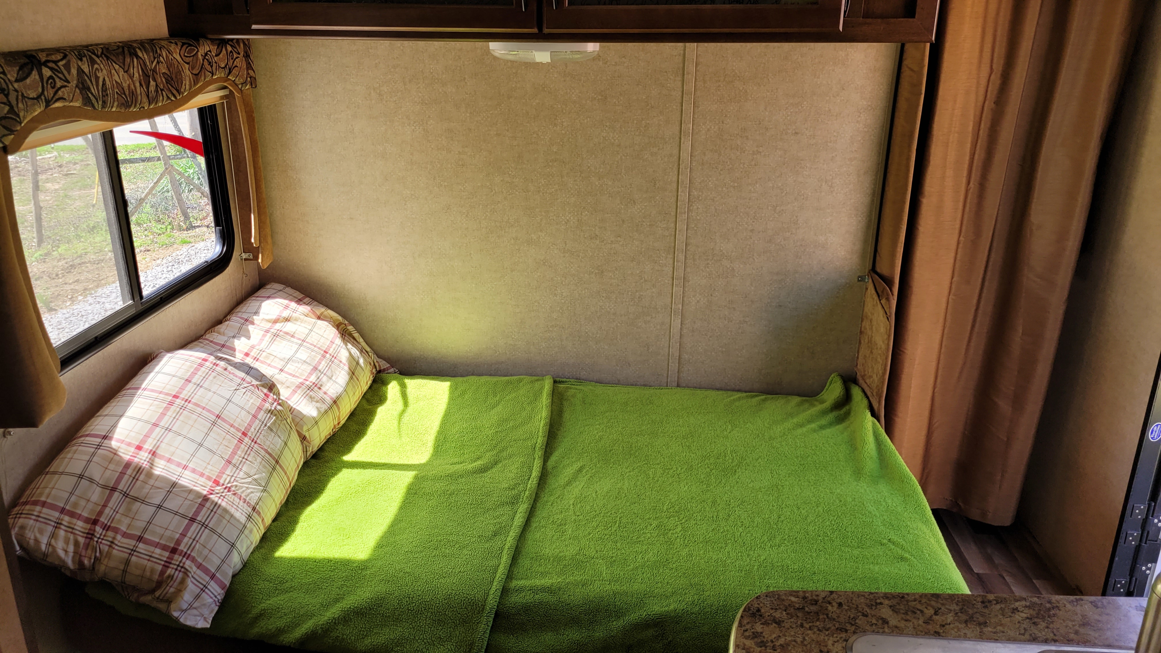 Cozy bed area in an RV rental, featuring a green blanket, plaid pillows, and sunlight streaming through the window, ideal for travel.. Pacific Coachworks Tango Widelite 2015