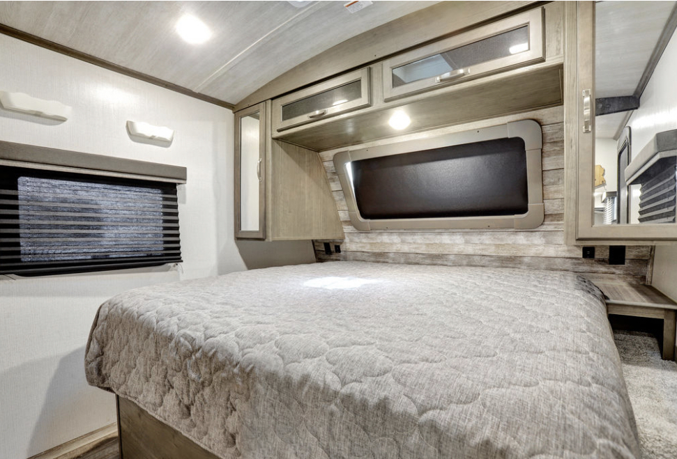 Inside an RV rental: a cozy bedroom with a large quilted bed, overhead cabinets, window blinds, and a built-in nightstand.. Keystone RV Cougar 2021