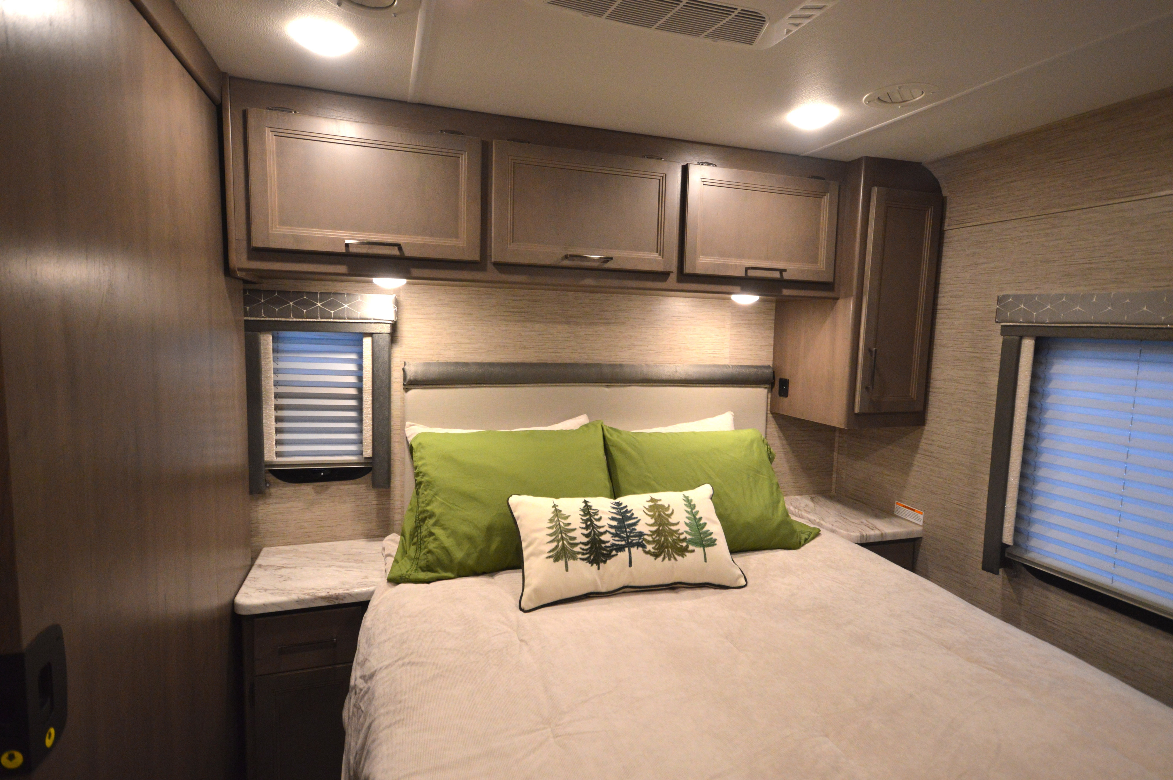 Cozy RV rental bedroom featuring a large bed with green and decorative pillows, overhead cabinets, and side windows.. Thor Motor Coach Freedom Traveler 2023