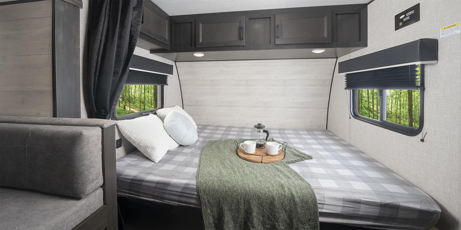 Cozy RV rental bedroom featuring a plaid bed, overhead cabinets, and scenic forest views from the windows.. Jayco slx 2023