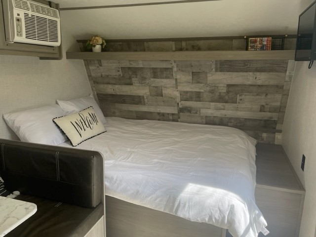 Comfortable RV rental sleeping area featuring white bedding, a 'WELCOME' pillow, wood accent wall, and AC.. Dutchmen Coleman Lantern LT 2023