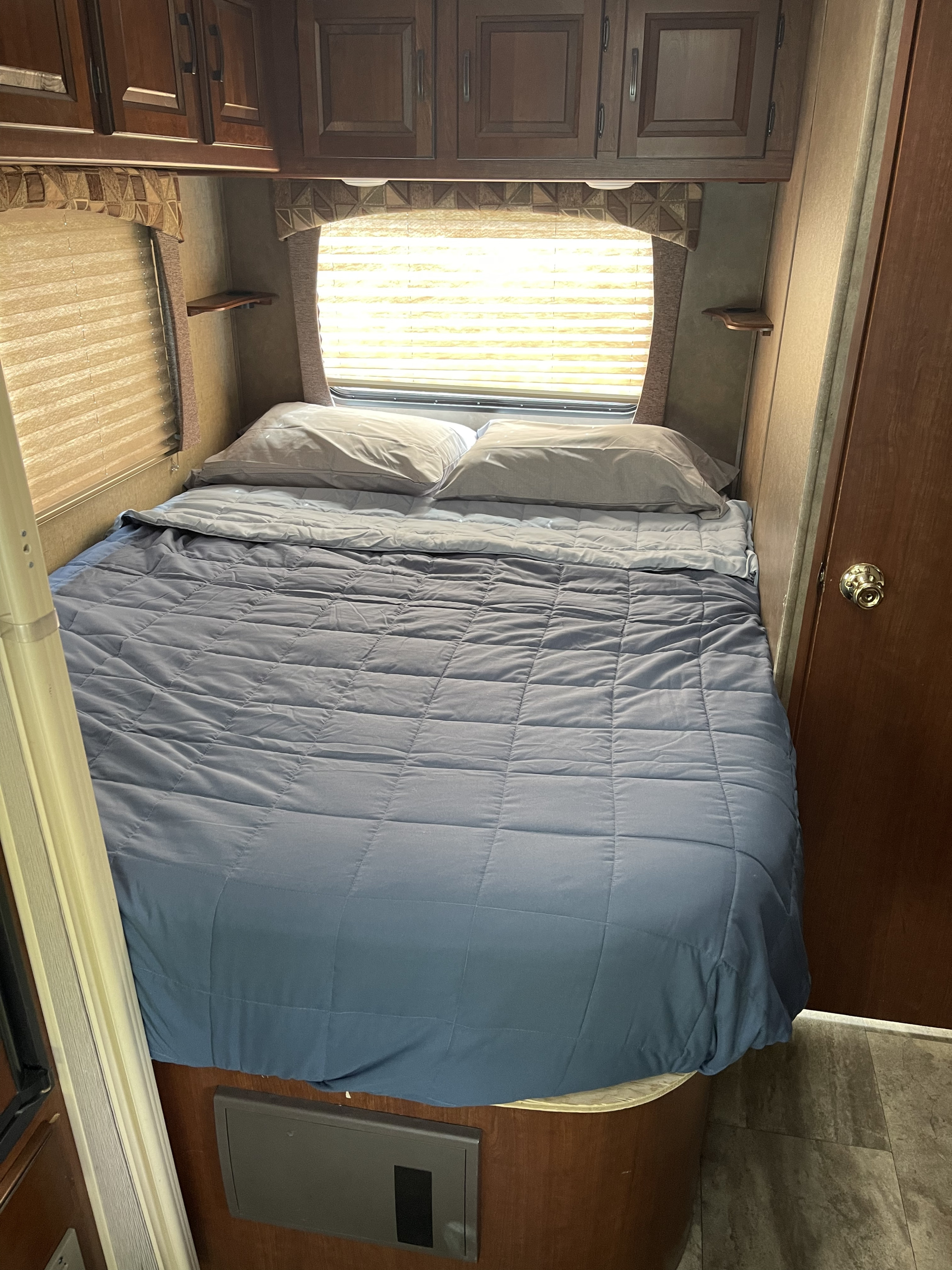 Comfortable RV rental bedroom featuring a made-up bed with blue comforter, pillows, overhead cabinets, and bright windows.. Forest River Sunseeker 2016