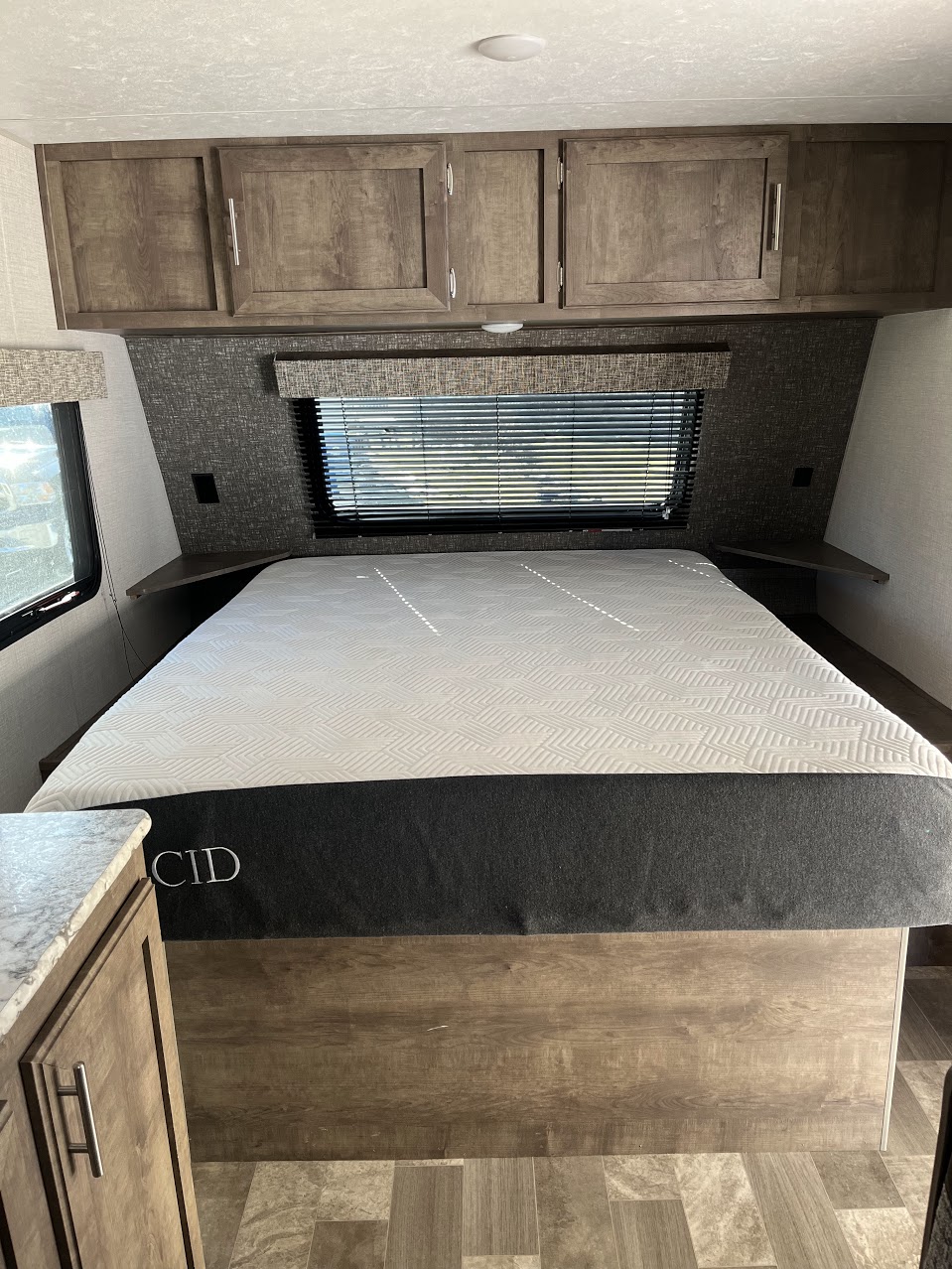 Compact RV rental bedroom showing a mattress, overhead cabinets, window with blinds, and small side shelves.. Sportsmen Classic 2020