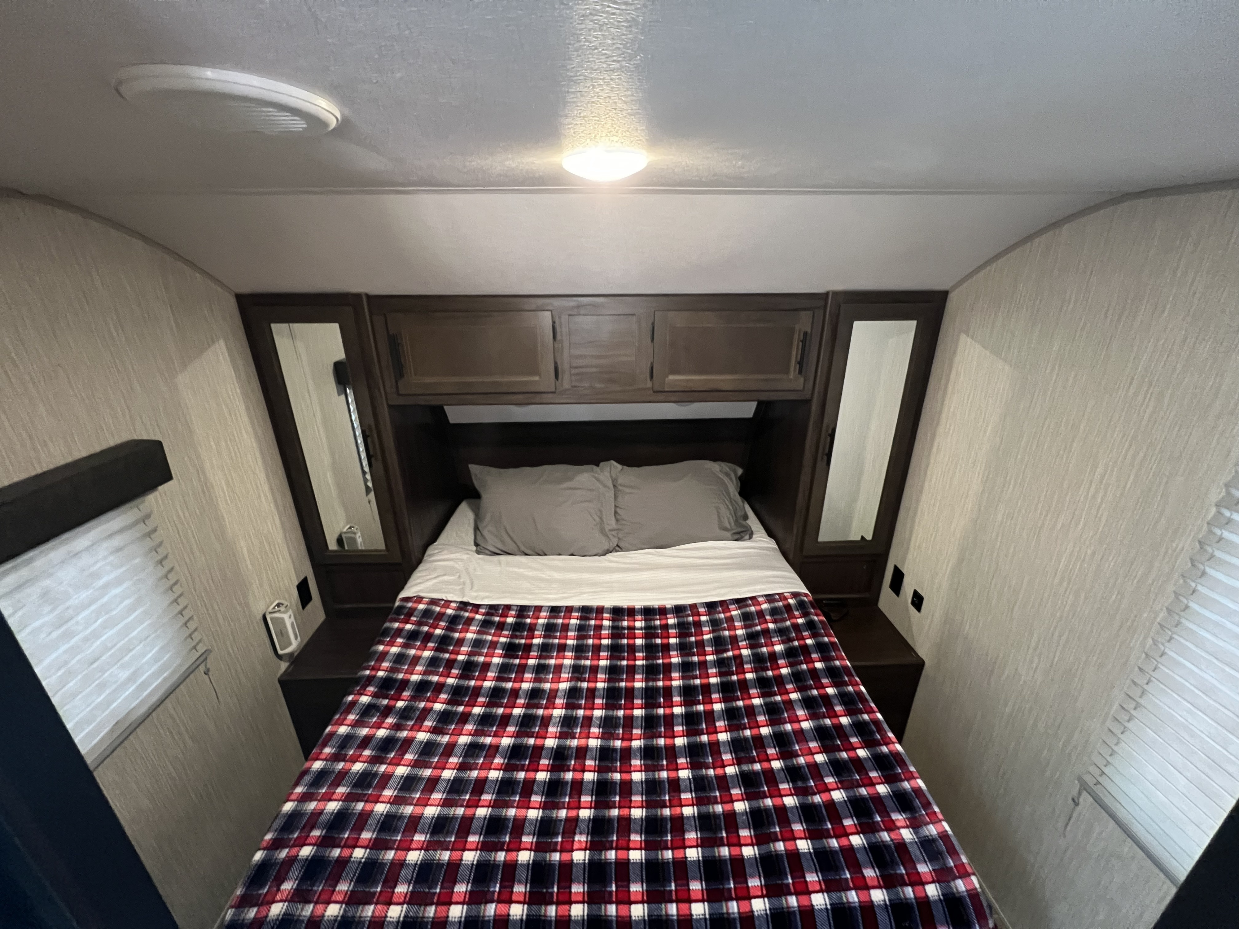 This inviting RV rental bedroom offers a comfortable bed with plaid blanket, overhead storage, side mirrors, and ceiling lights.. Forest River Cherokee Grey Wolf 2020