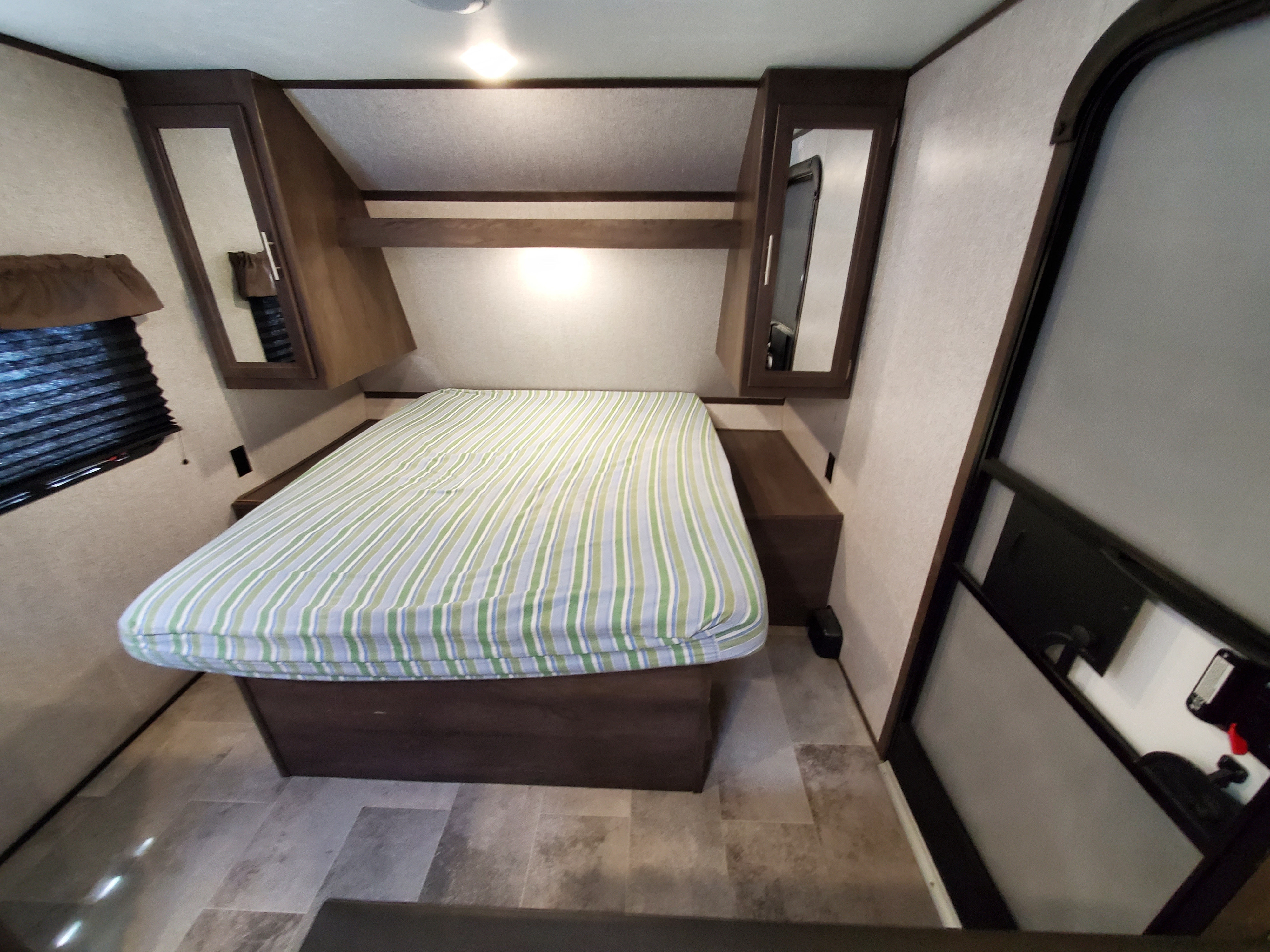 Cozy rv rental bedroom features a striped bed, mirrored overhead cabinets, and a window with blinds. Ready for your trip!. Dutchmen Coleman 2020