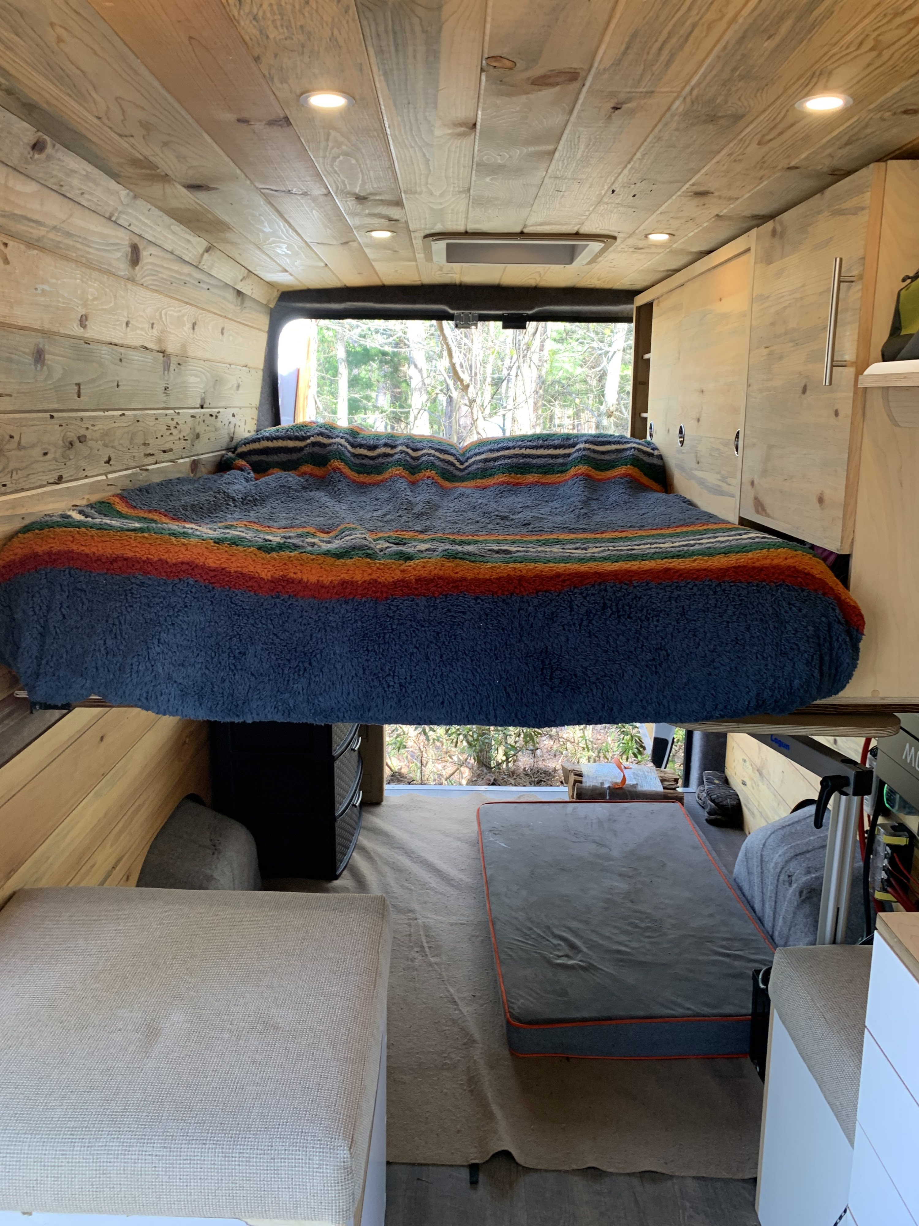 Cozy camper van interior for RV rental. Features a raised bed with striped blanket, wood paneling, and storage. Ideal for travel adventures.. Dodge Sprinter Van 2020