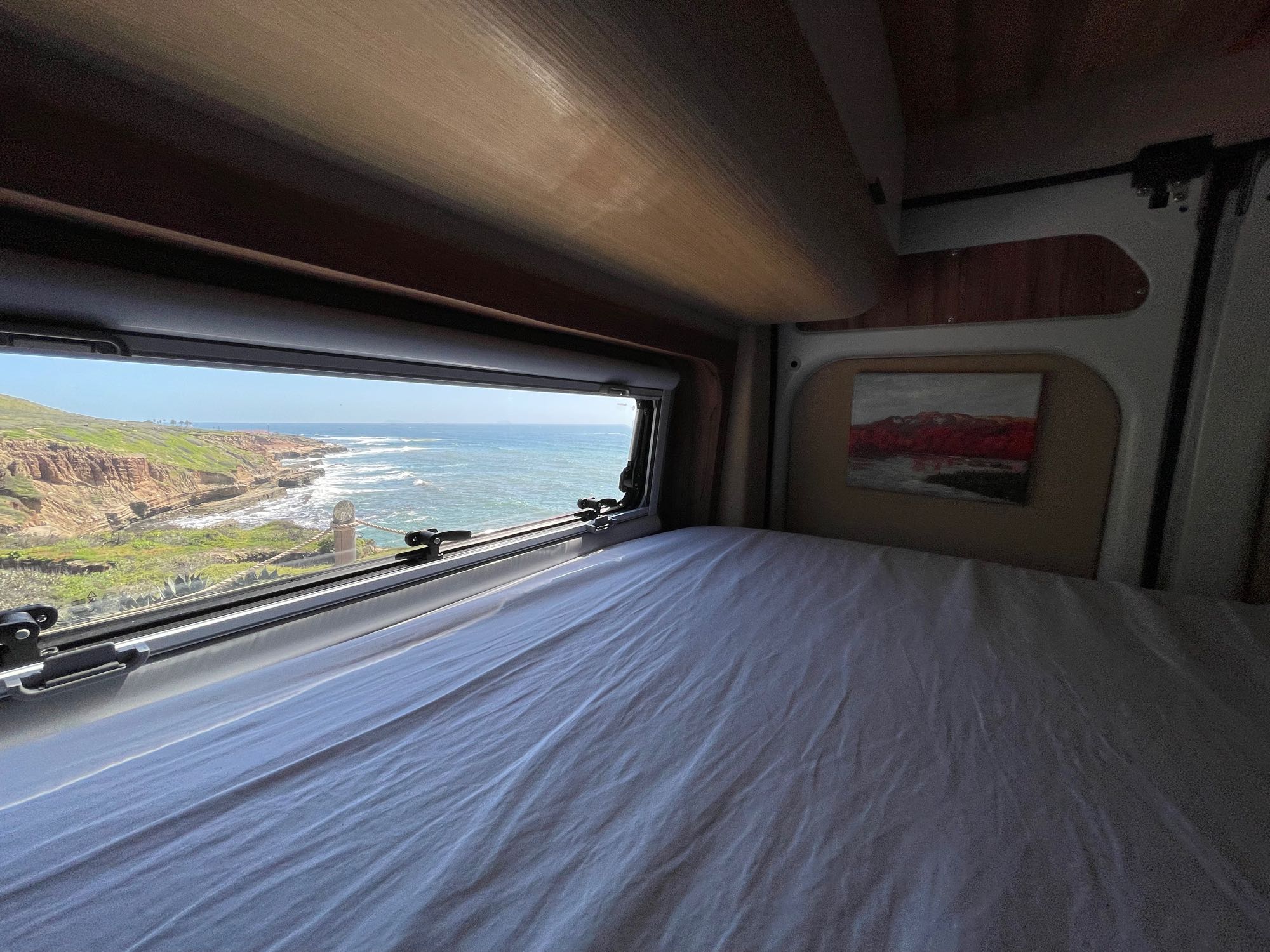 Inside an RV rental, a bed faces a large window revealing a stunning view of a rocky coastline and ocean waves.. BEMYVAN B-Adventure 2022