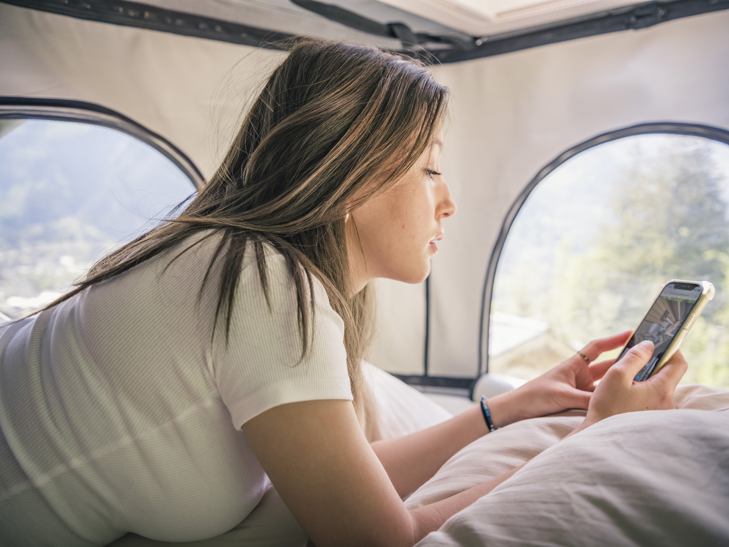 Young woman relaxing in an RV rental, browsing her phone in bed with scenic views out the windows.. Fiat Ducato L2H2 2020