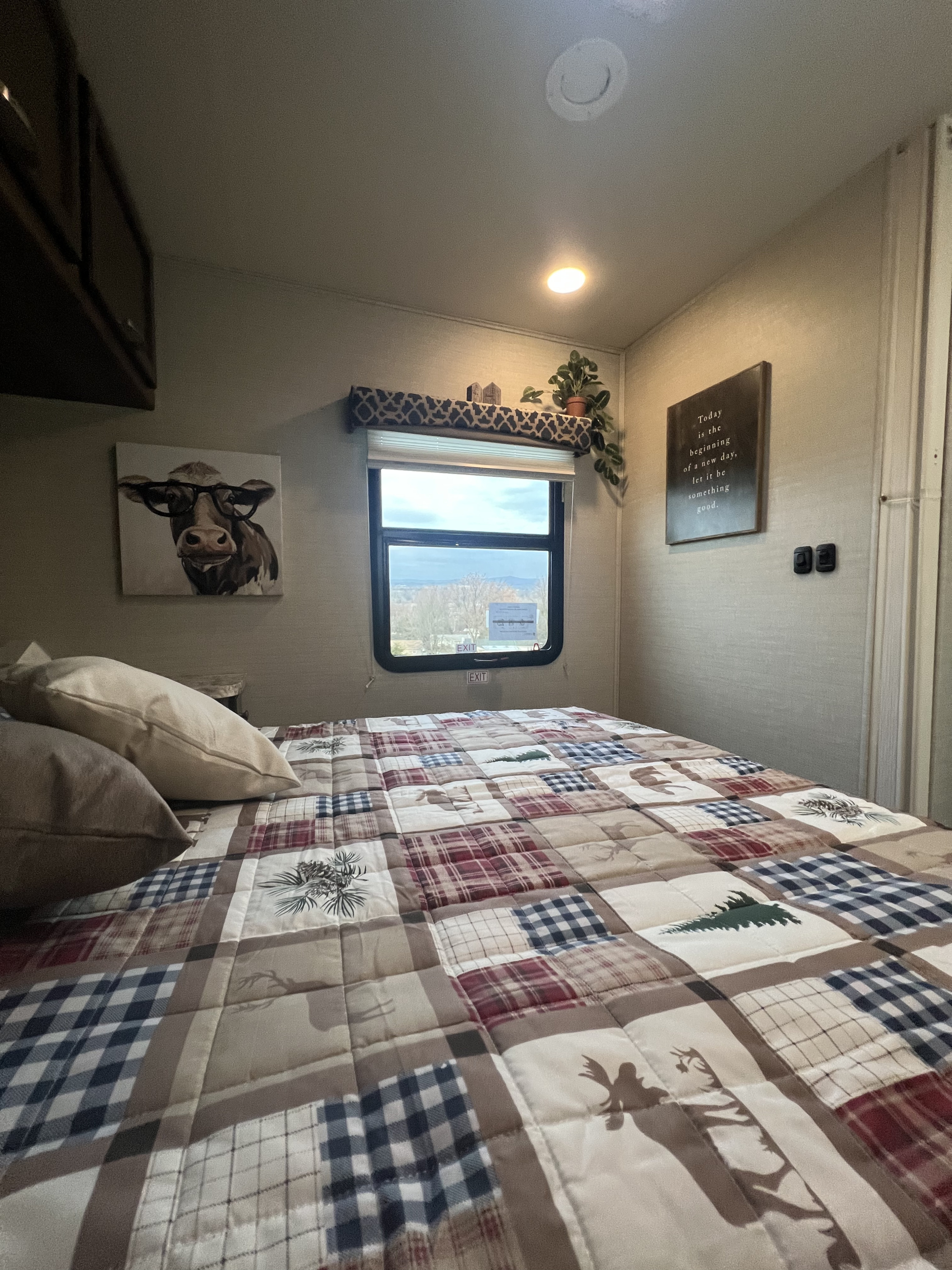 Cozy RV rental bedroom features a rustic plaid quilt, cow art, and a scenic window view for a restful stay.. Jayco Redhawk 2019