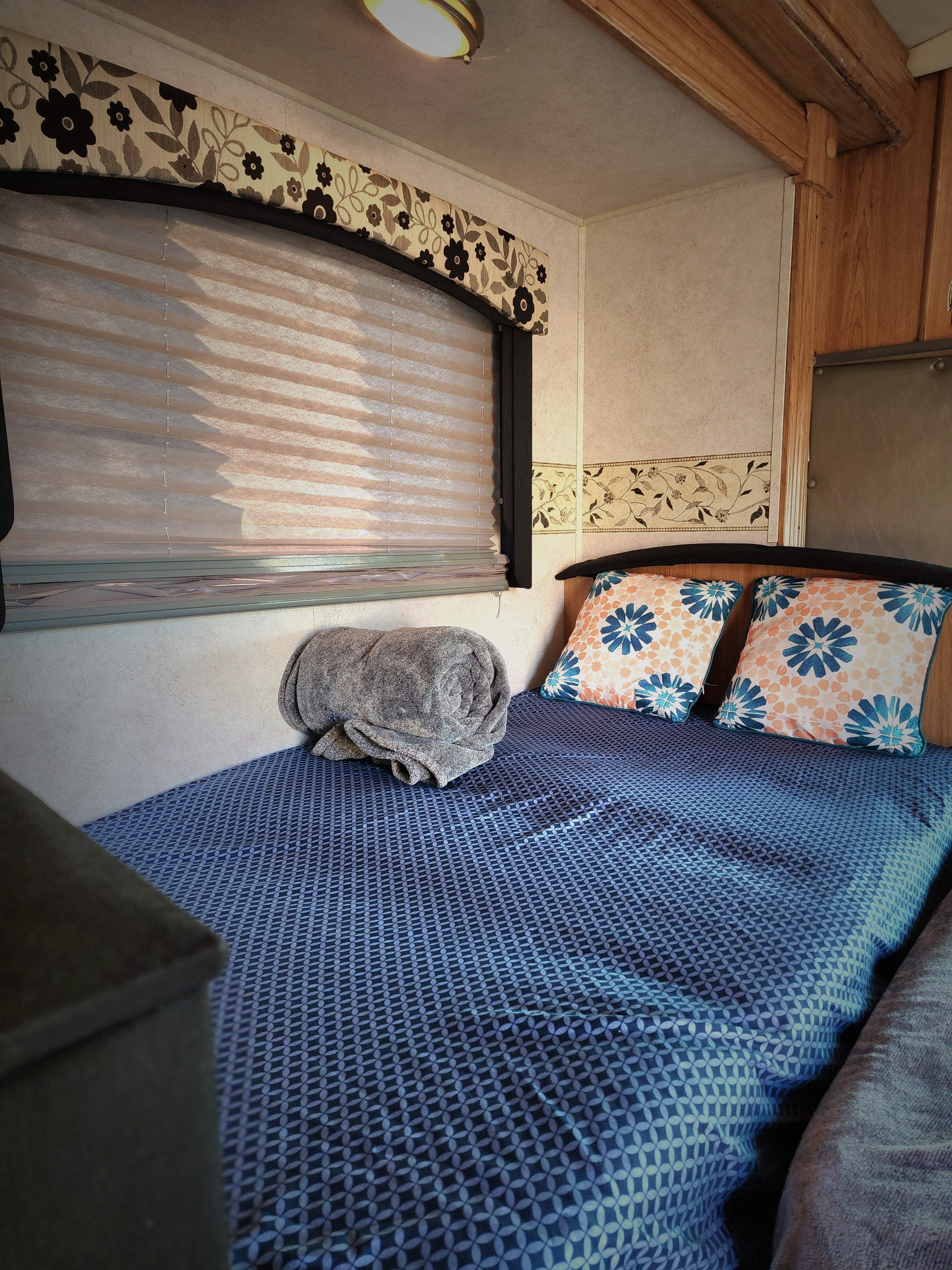 Cozy RV rental bedroom features a comfy bed with blue patterned bedding, floral pillows, and a window with blinds and valance.. R-Vision Trail-Lite B-Plus 2006