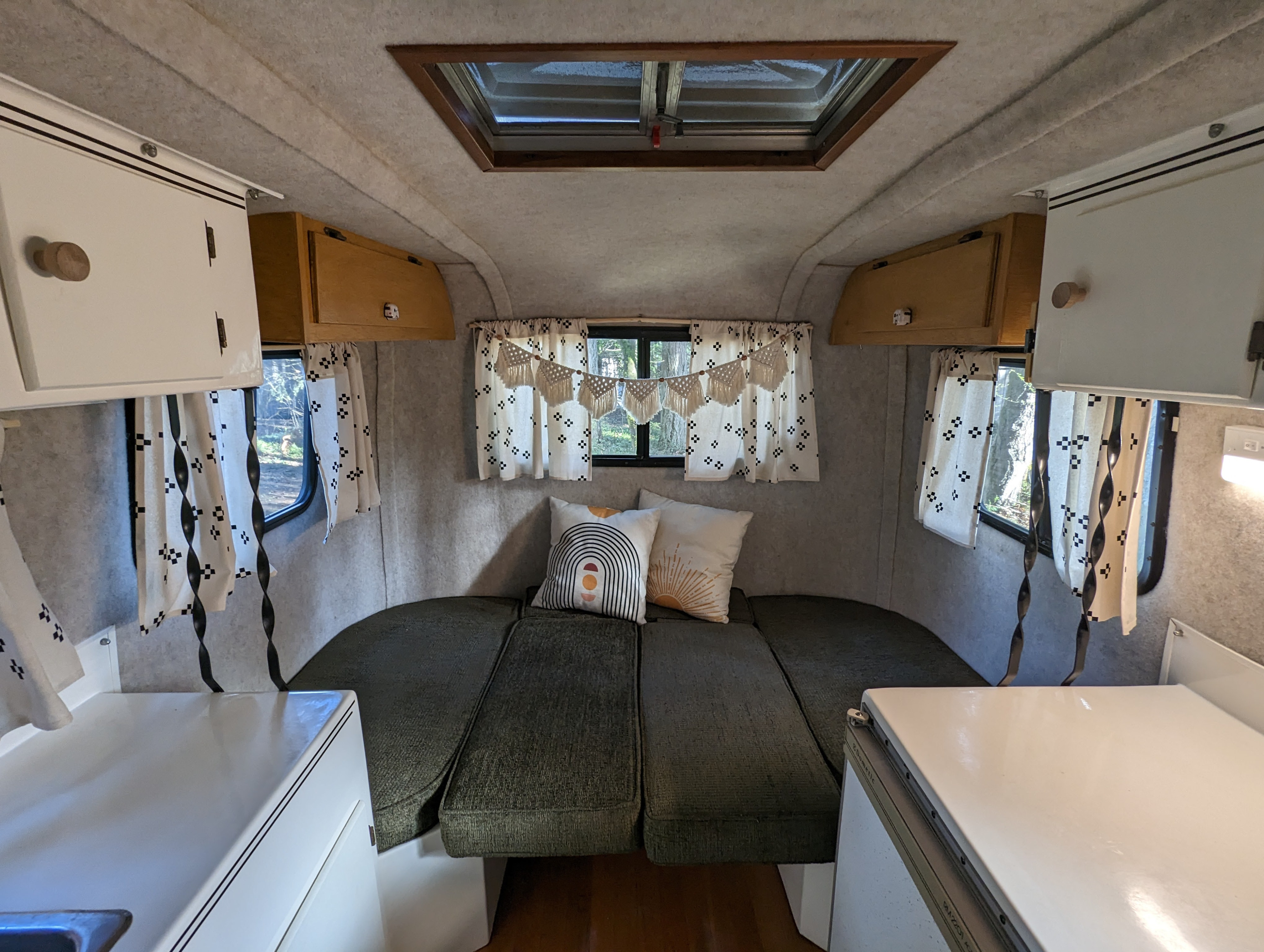 RV rental interior view: cozy bed/seating area, white kitchen counters, and windows with decorative curtains.. Scamp 16' 1992