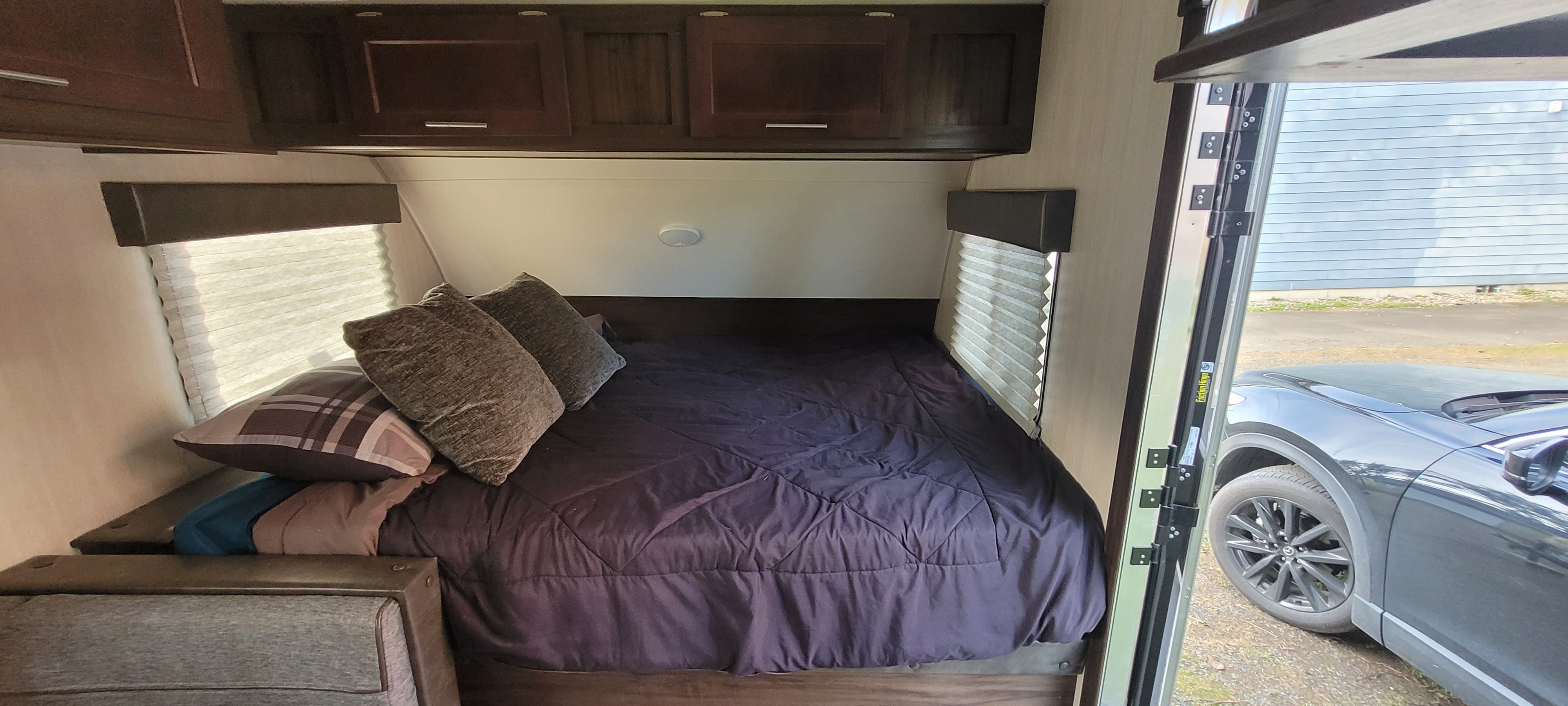Cozy RV rental bedroom with dark bedding, overhead cabinets, and window blinds. A car is visible outside.. Forest River Cascade 2019
