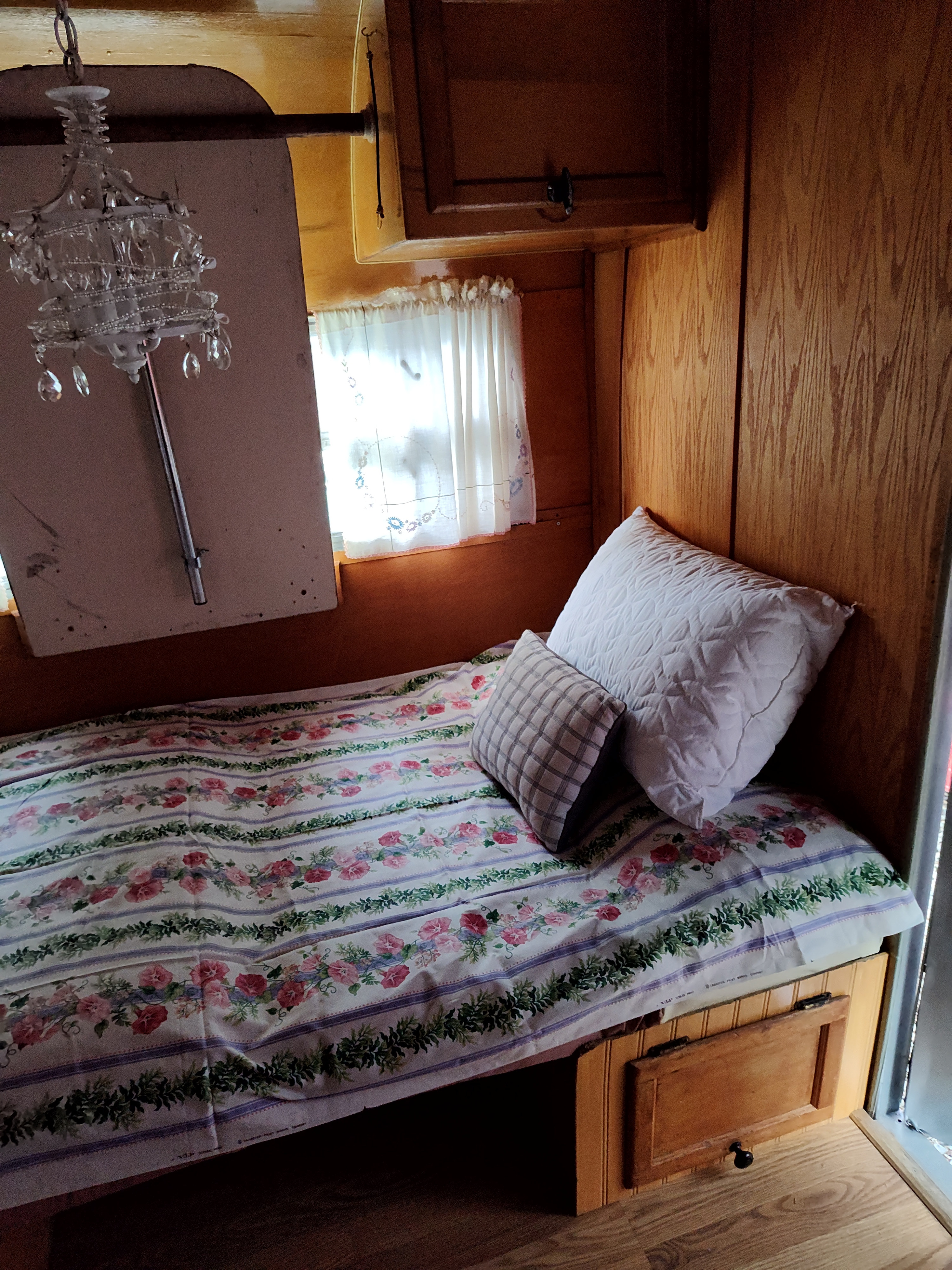 RV rental bedroom: cozy bed with floral bedding & pillows, wooden paneling, window with curtains, and a chandelier.. Nomad camp trailer 1957