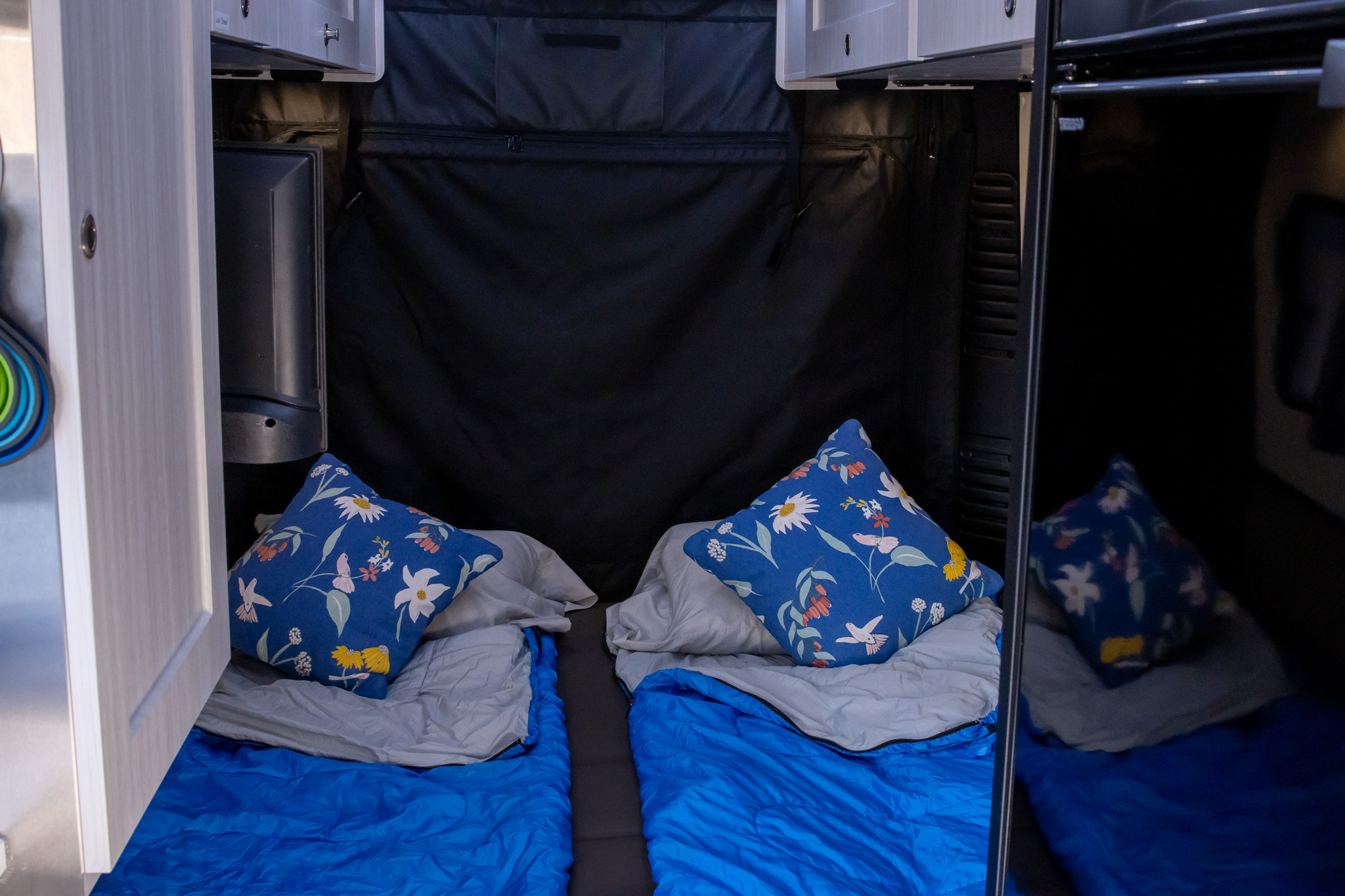 Cozy RV rental interior with two beds, blue sleeping bags, floral pillows, plus a mounted TV and shiny fridge panel.. RoadTrek Zion Slumber 2023