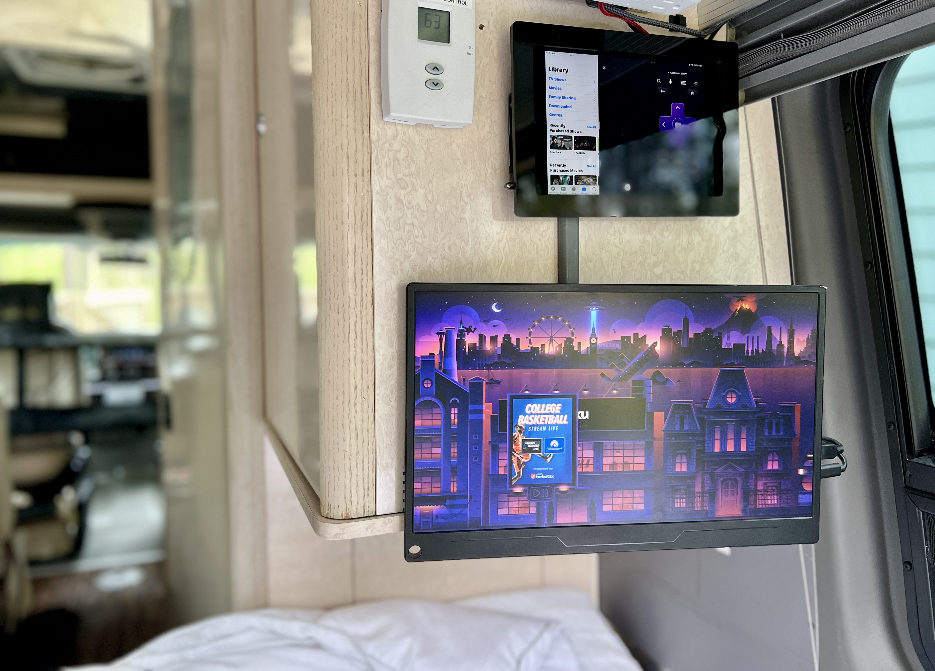 RV rental interior: Two wall-mounted screens with a media library tablet and a monitor showing a college basketball ad; thermostat visible.. Airstream Interstate 2010