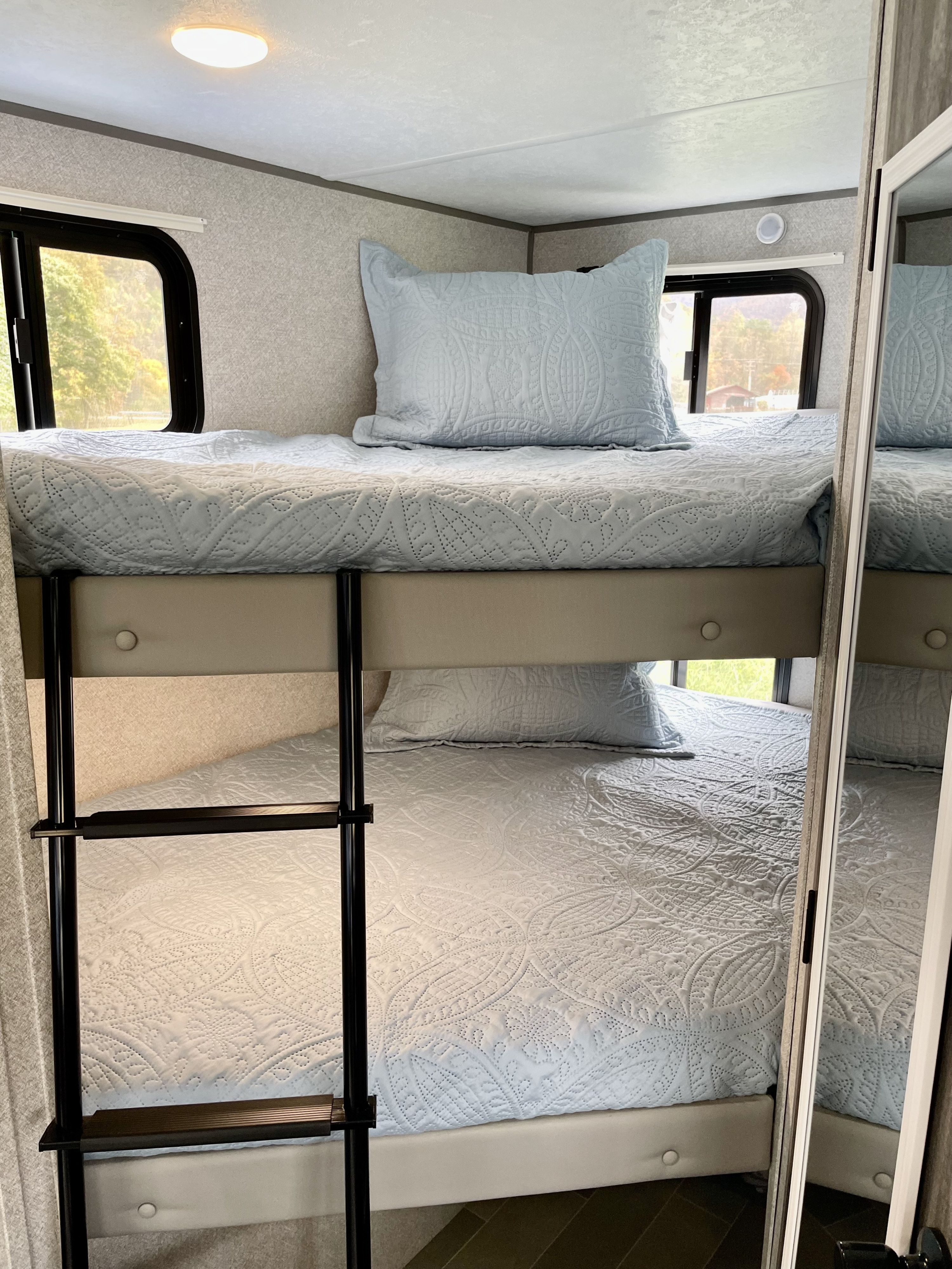 A bright RV rental interior featuring stacked bunk beds with soft blue quilted bedding and a ladder. Windows show outdoor scenery.. Forest River Salem FSX PLATINUM 2020