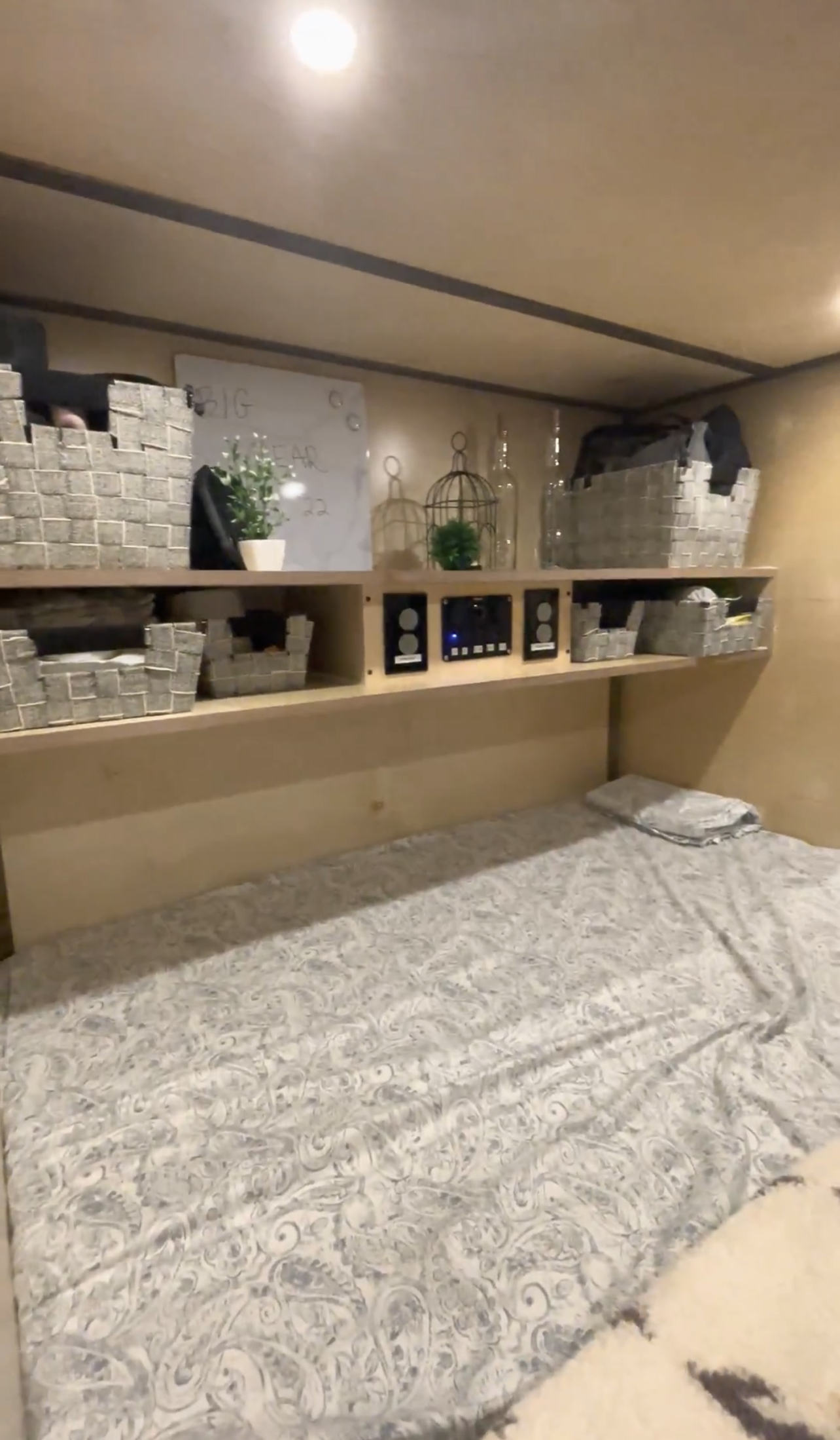 Cozy RV rental interior shows a bed with patterned sheets, built-in wooden shelving with storage baskets, and control panels.. Pura Vida Overland PV1 2022