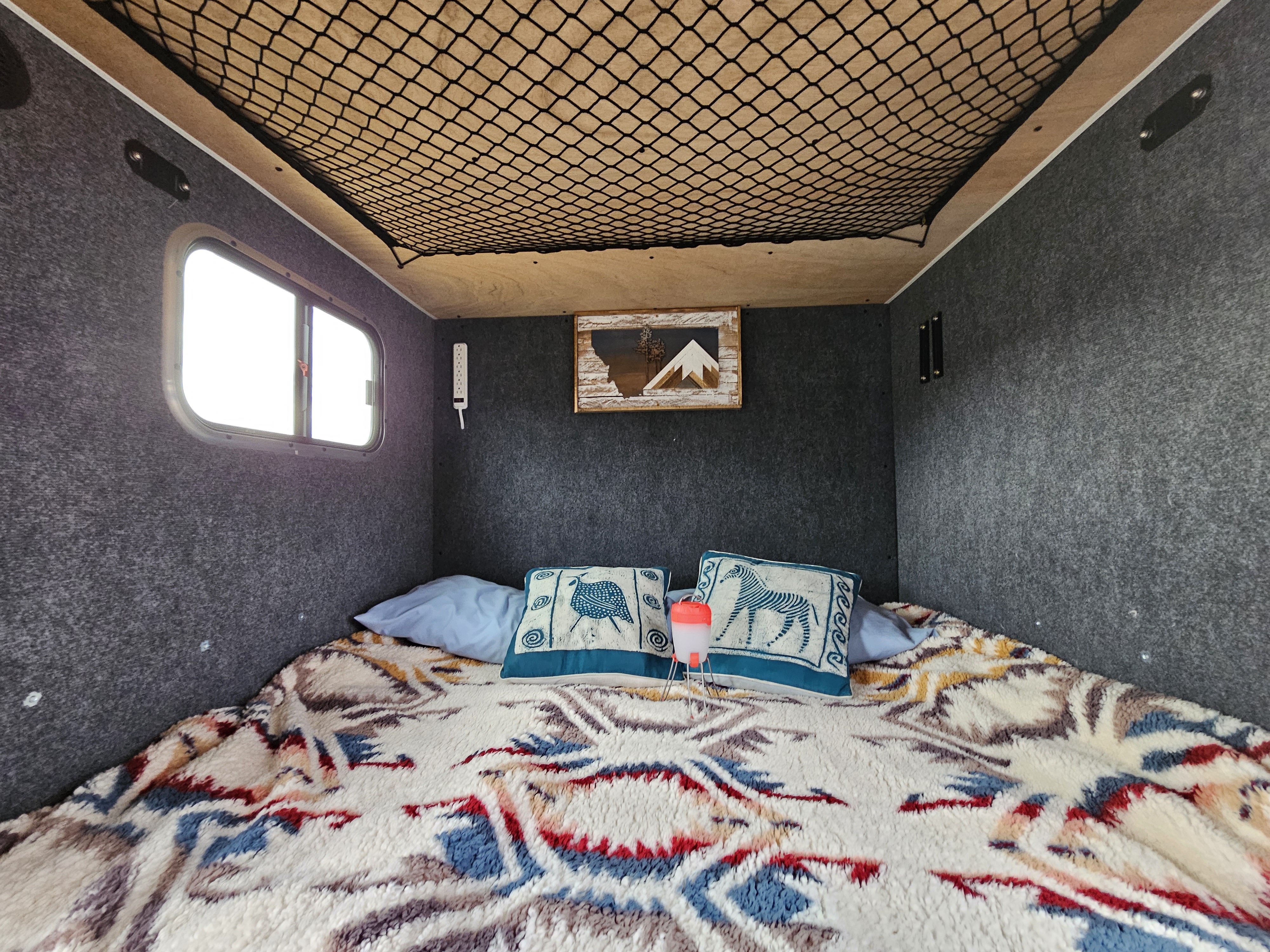 Explore this cozy RV rental interior featuring a comfy bed with patterned blanket, soft gray walls, window, and overhead storage net.. Sherpa Big Foot 2020